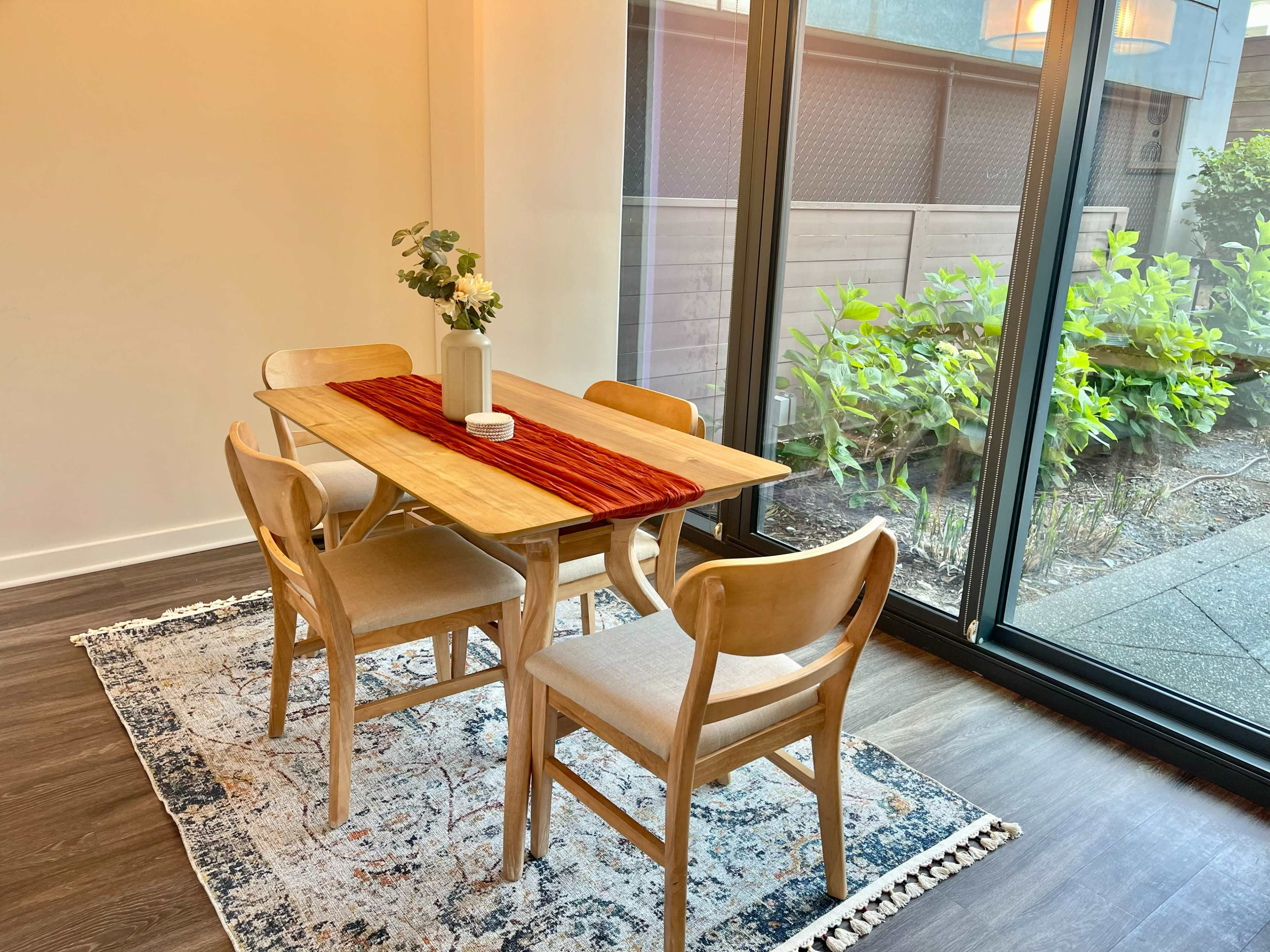 A wooden dining table with four chairs is set near a large glass window overlooking a garden.