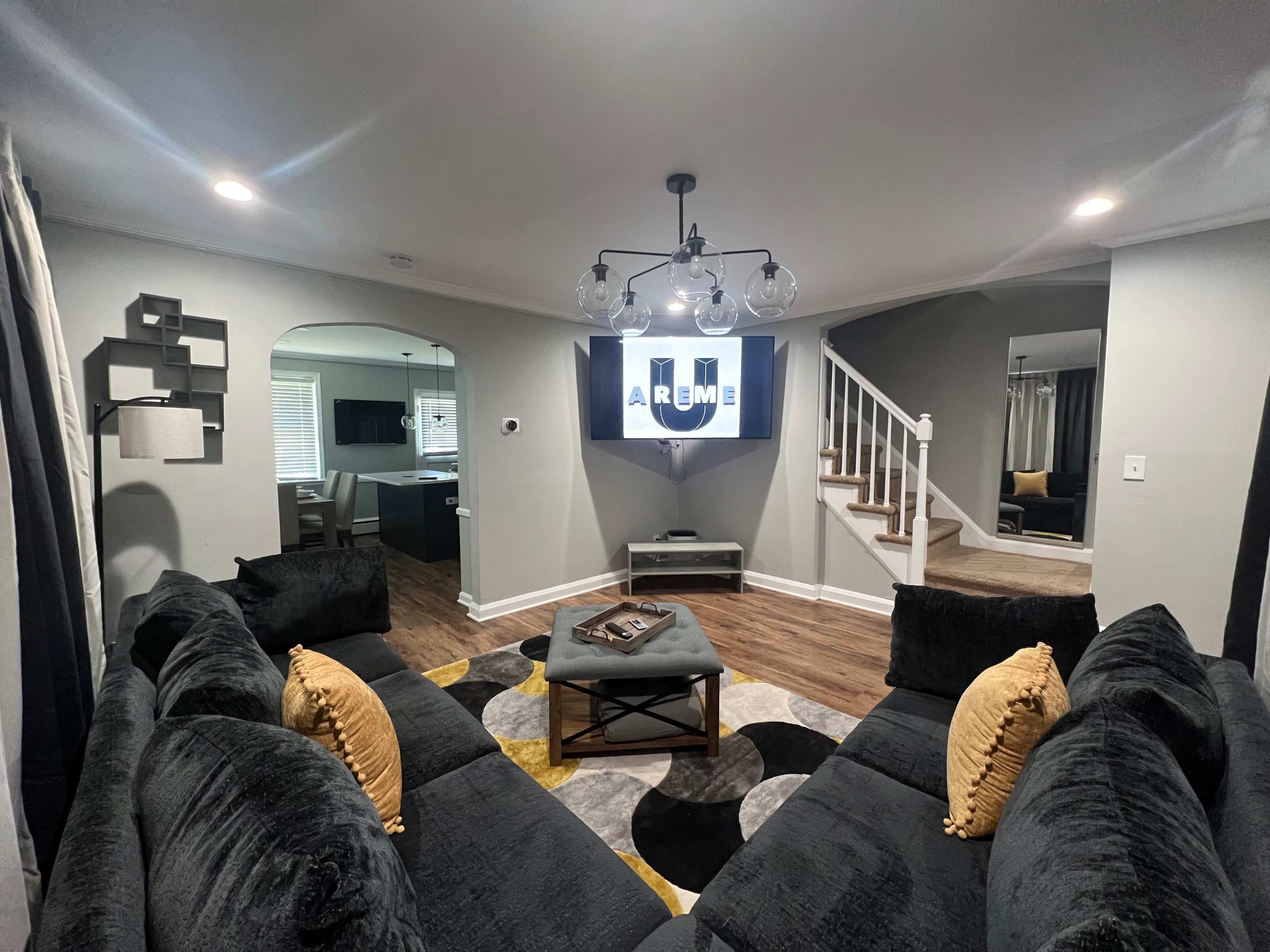 The living room features a black sectional sofa arranged around a circular patterned rug, with a modern chandelier overhead and a TV mounted on the wall.