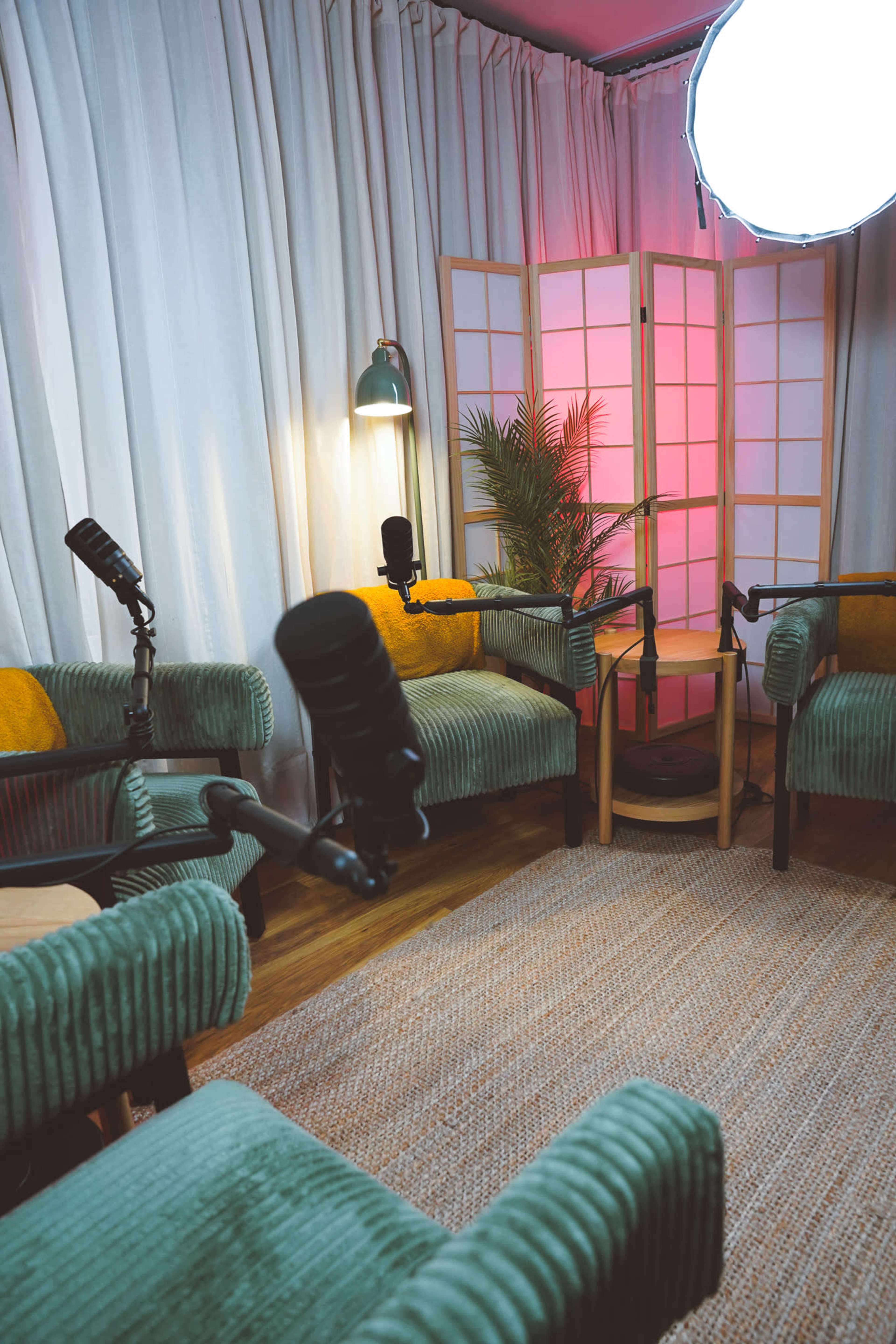 The image shows a cozy podcasting setup featuring green armchairs, microphones on stands, a plant, a lamp, and a lighted screen in the background.