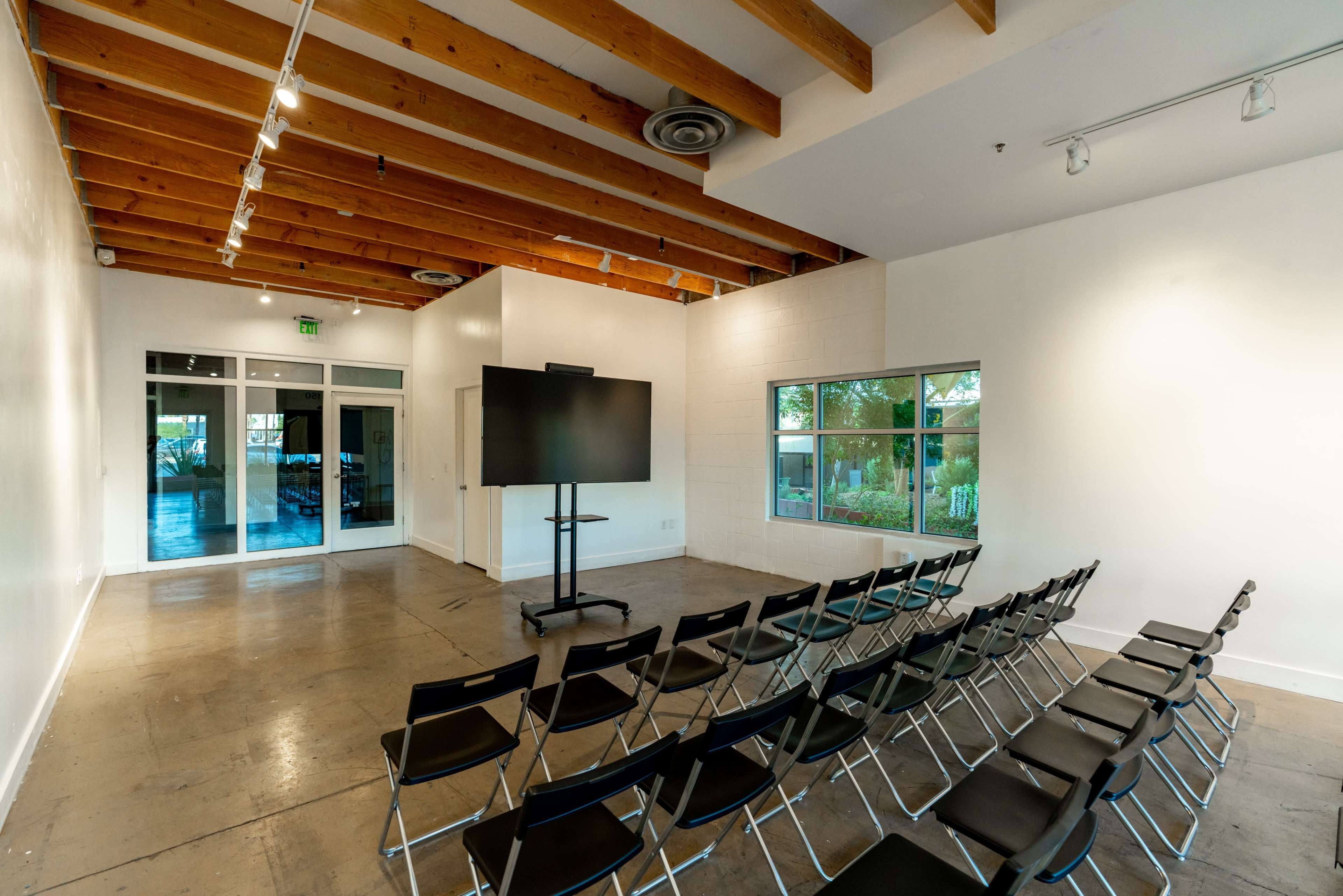 A modern meeting room with a row of black chairs facing a large television screen on a stand, set against a bright, minimalistic backdrop.