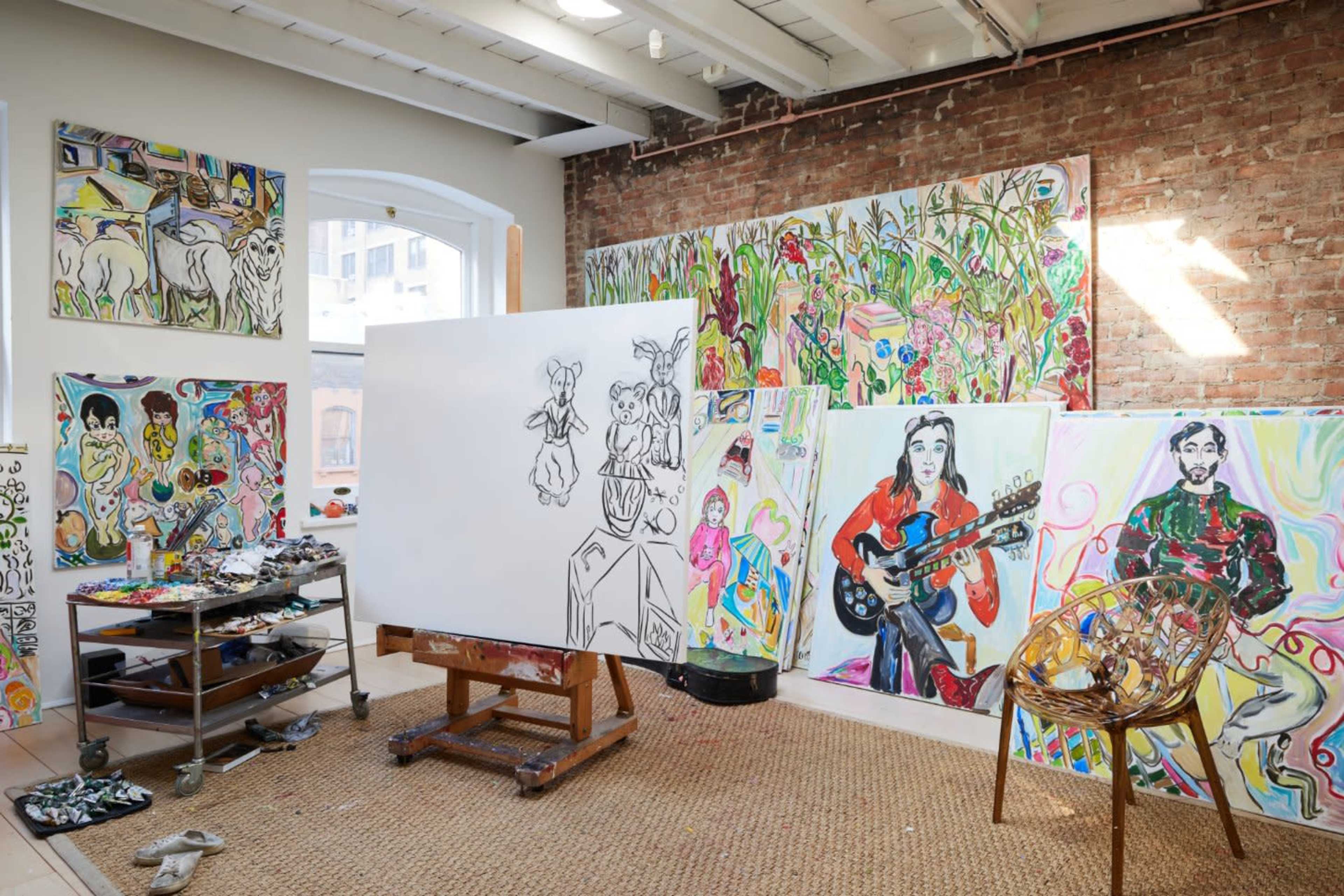 The image shows an art studio with various colorful paintings and sketches displayed on brick walls, a large blank canvas on an easel, and an artist's palette with brushes on a nearby cart.