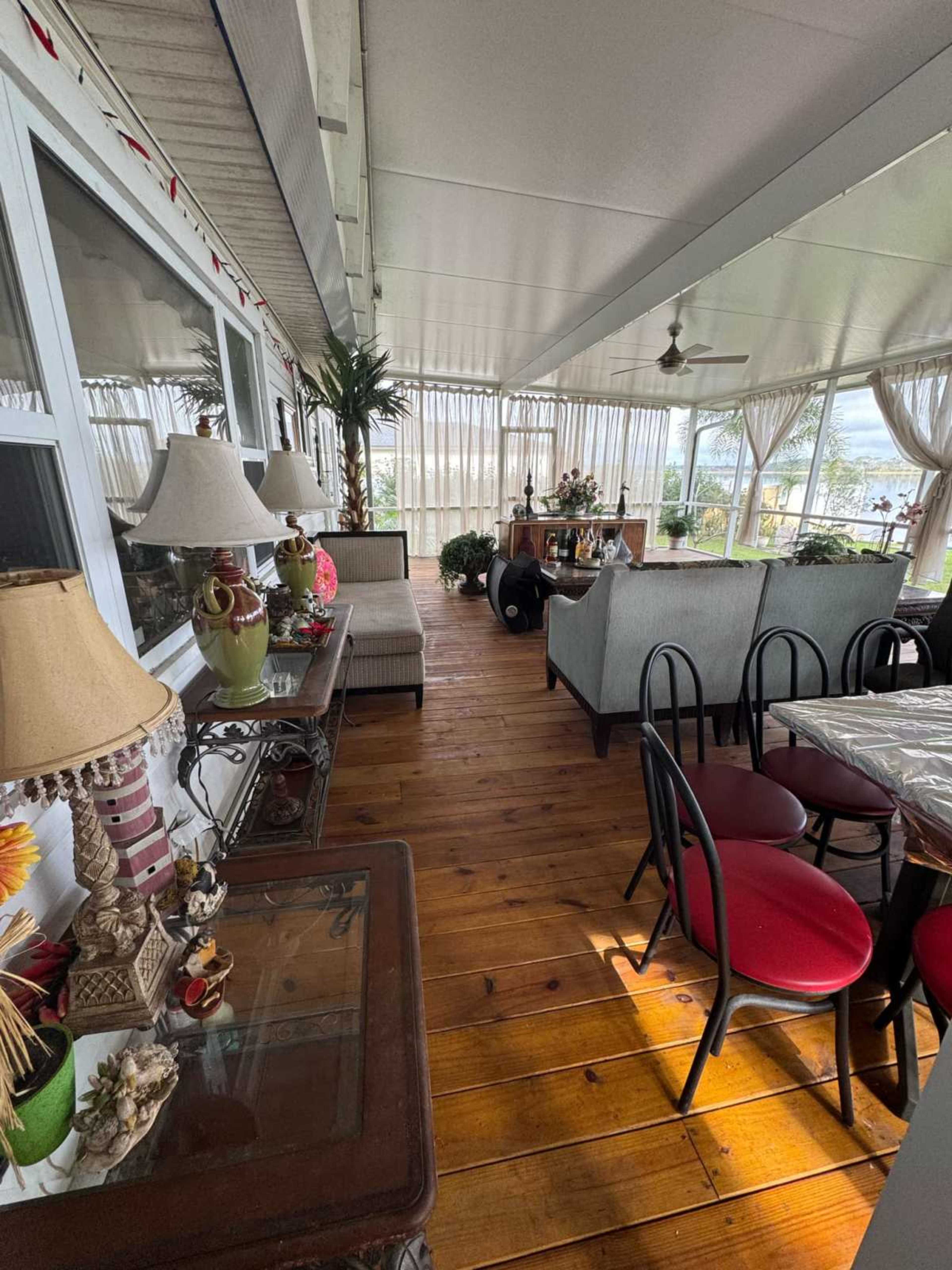 The image shows a sunlit enclosed porch featuring various plants, furniture, and decorative lamps arranged on wooden flooring.