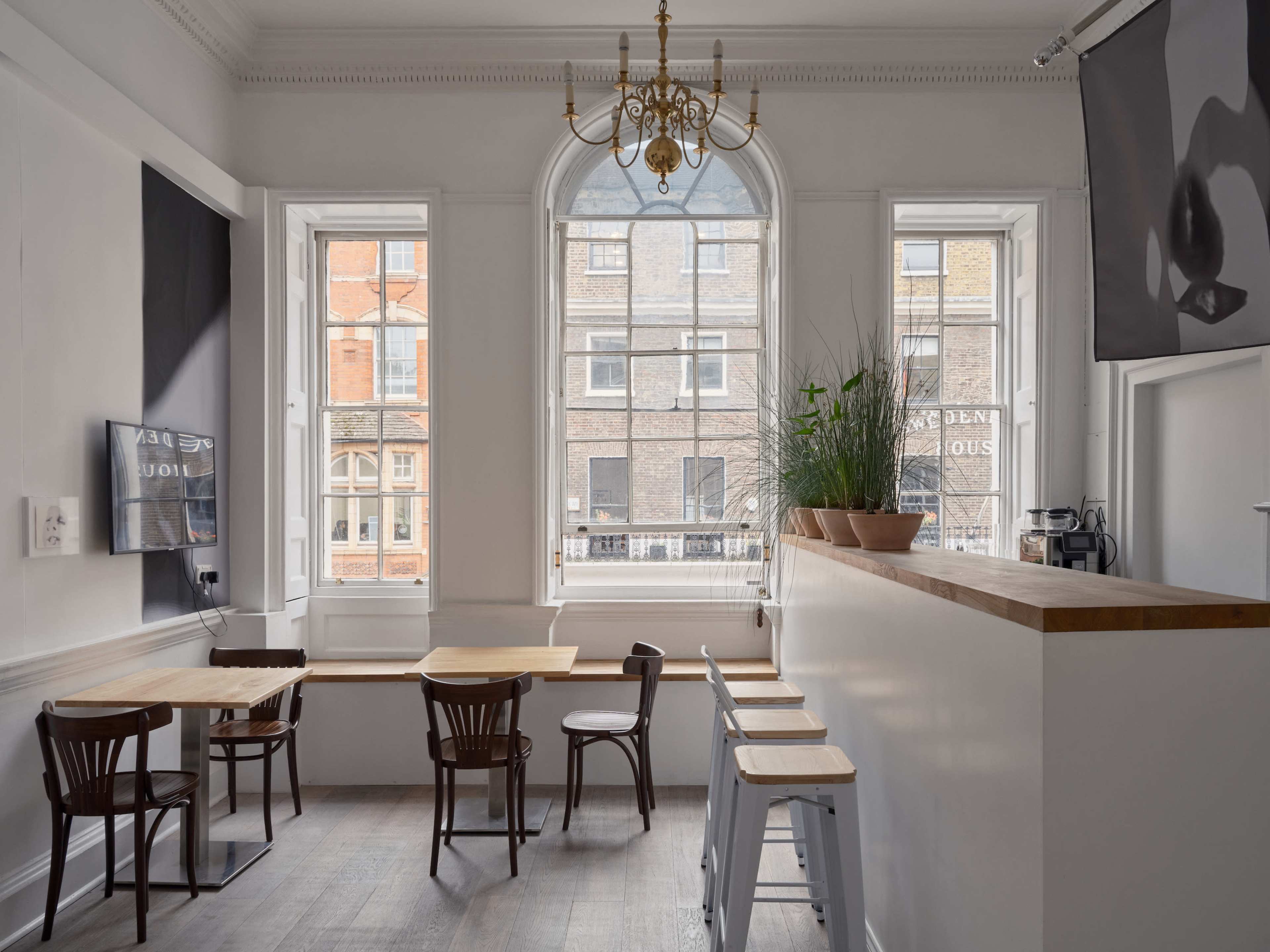 A modern cafe interior with wooden tables and stools, large windows allowing natural light, and a plant on the counter.