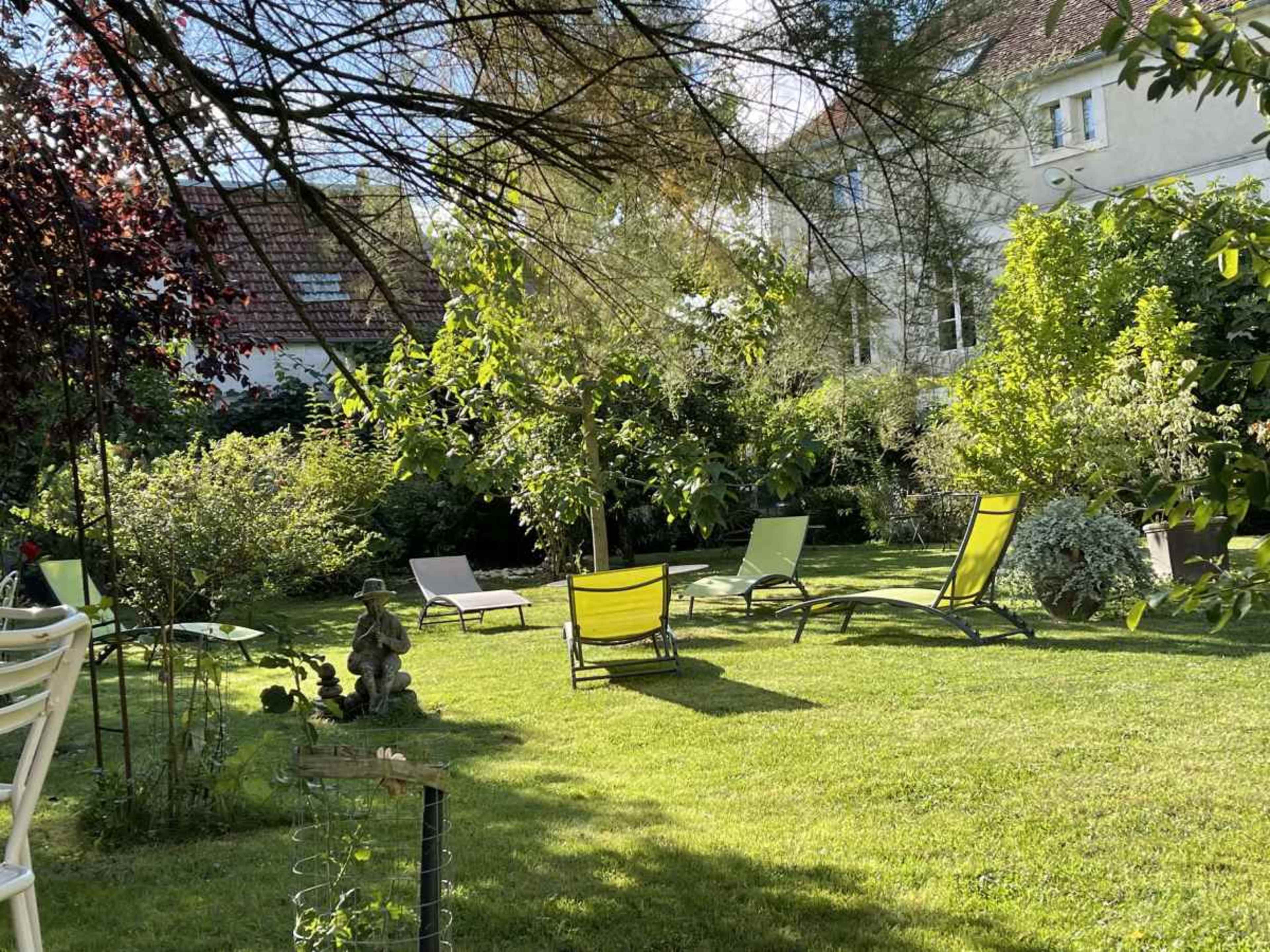 A tranquil garden scene features several lounge chairs positioned on green grass surrounded by lush foliage and trees.