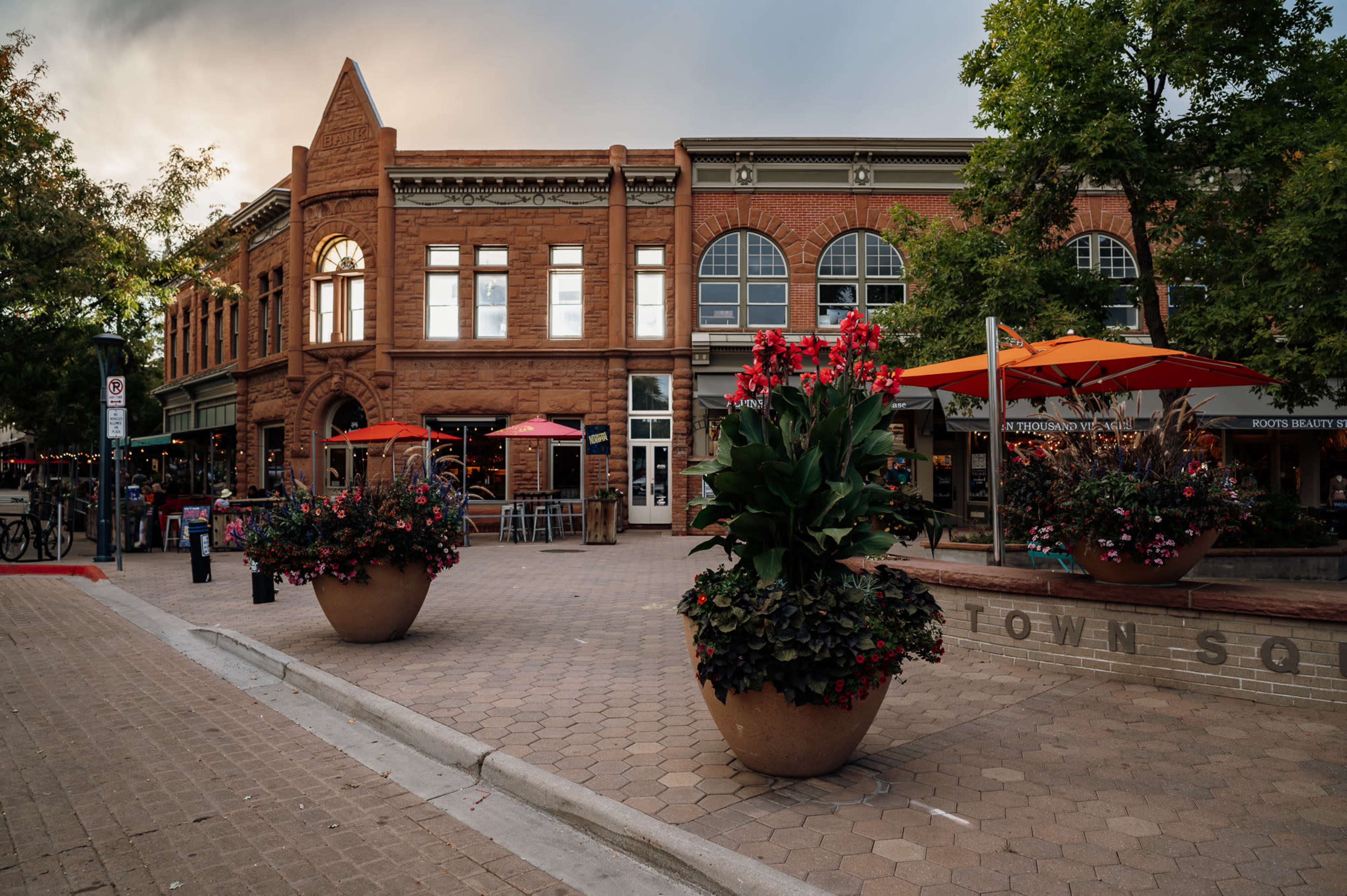 A pedestrian square lined with outdoor seating, vibrant flower pots, and historic brick buildings under a cloudy sky.