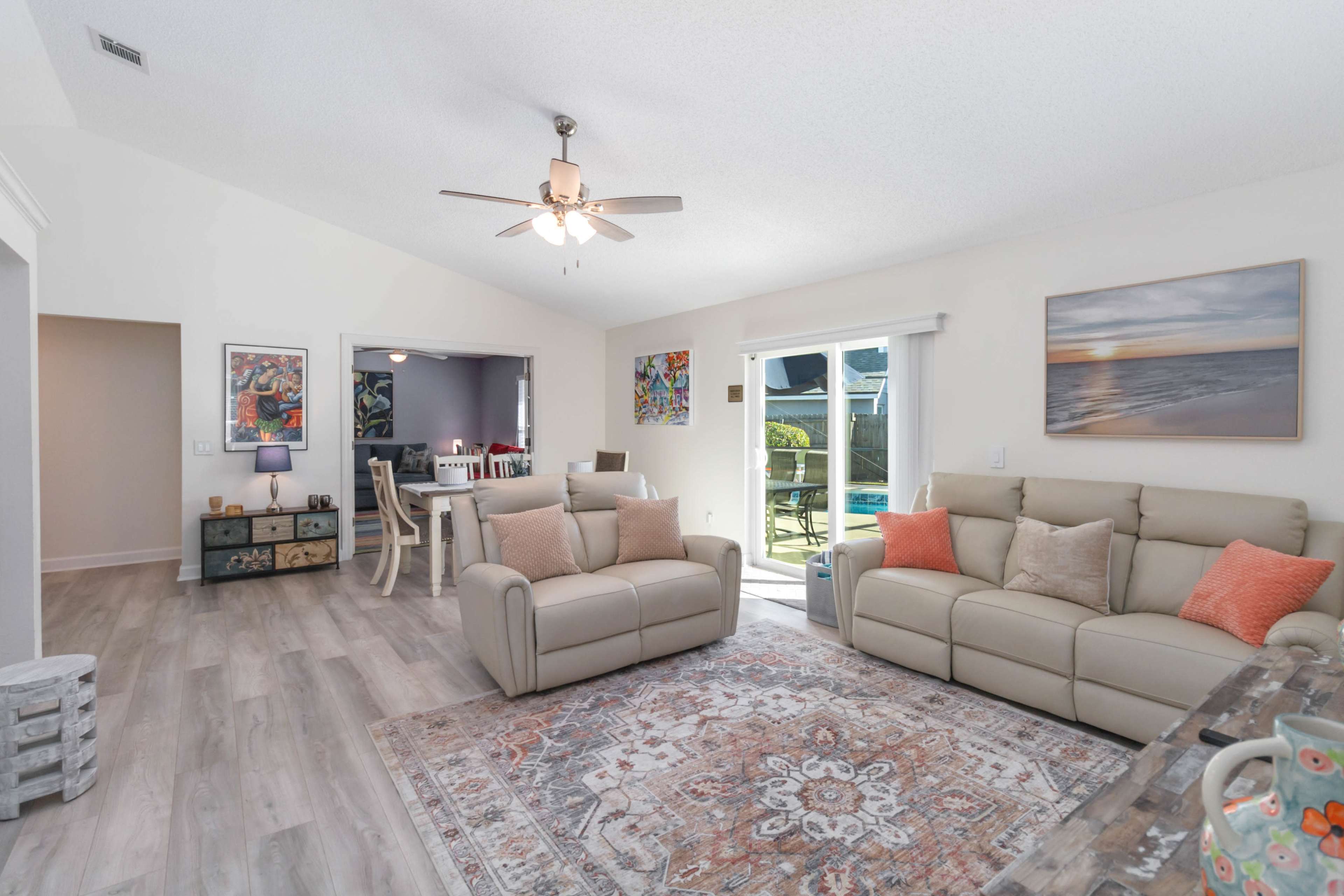 The living room features a beige couch, patterned area rug, and a dining area with a table and chairs, all set against light-colored walls and a ceiling fan.