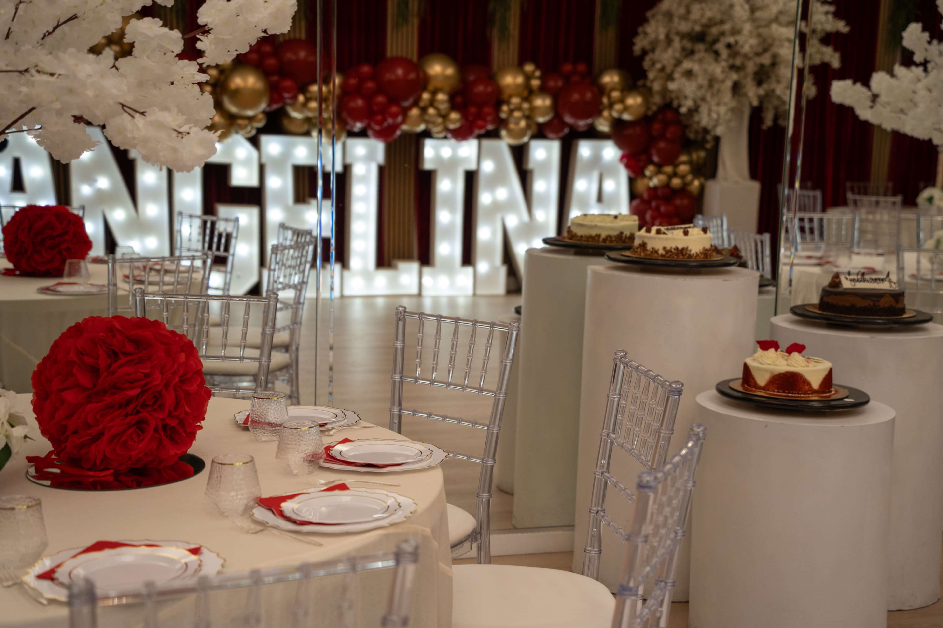 The image shows a elegantly decorated event space featuring white tables with floral centerpieces, illuminated letters spelling "ANGELINA," and various cakes displayed on pedestal tables.