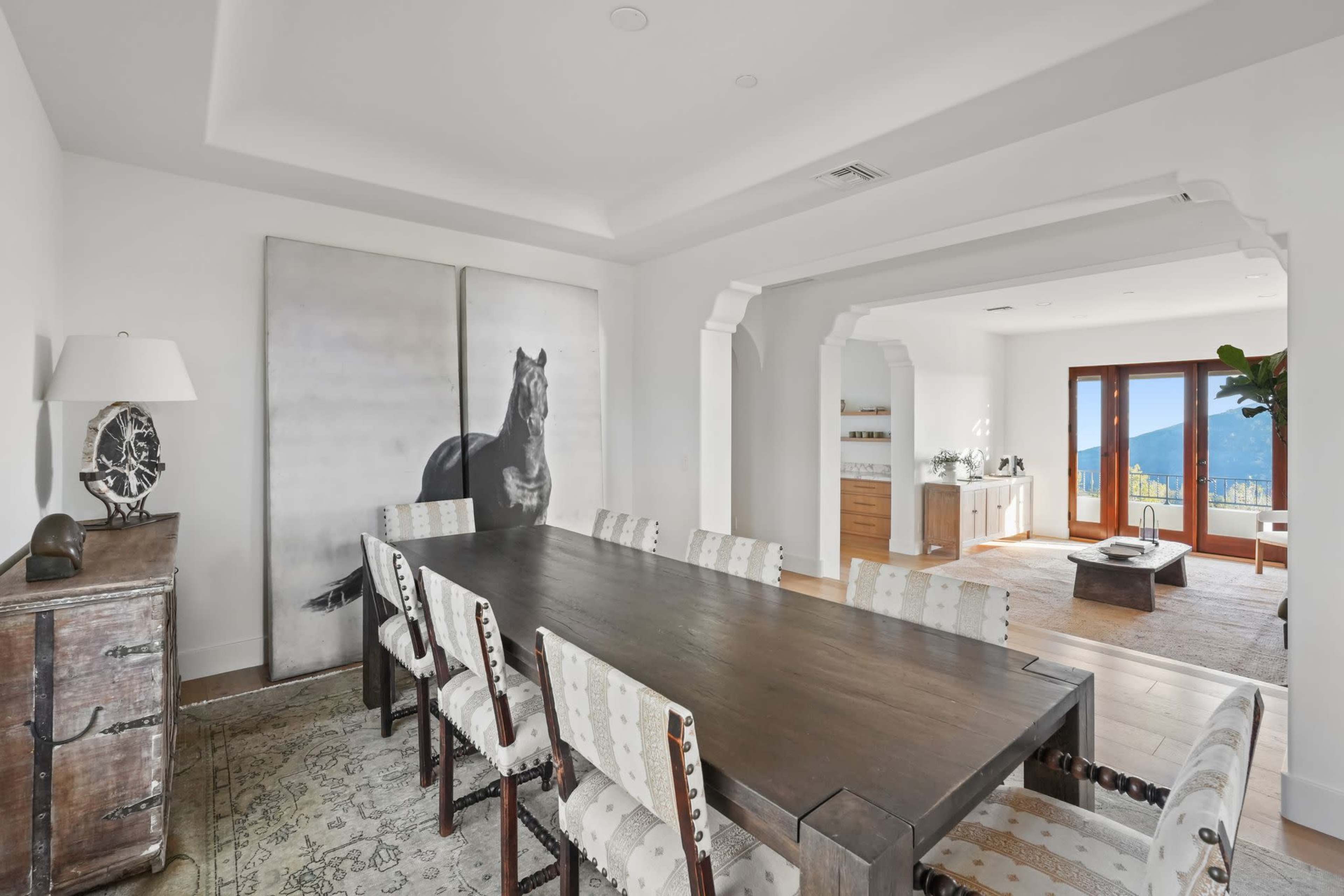 A modern dining room features a long wooden table surrounded by upholstered chairs, with a large horse painting on the wall and a view of mountains through glass doors.