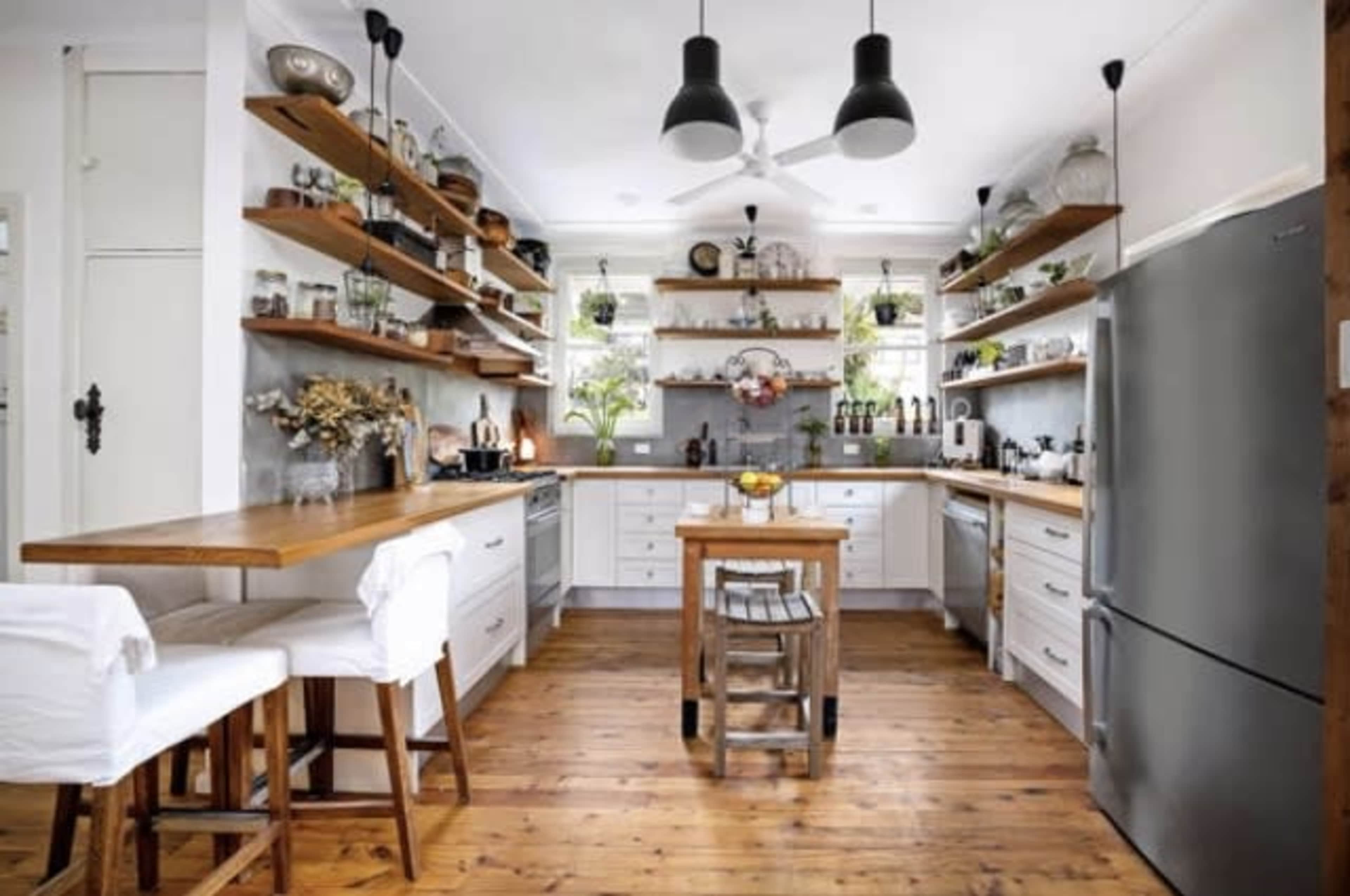 The kitchen features white cabinets, wooden shelves with various plants and decor, and a central table with chairs, all set on a wooden floor.