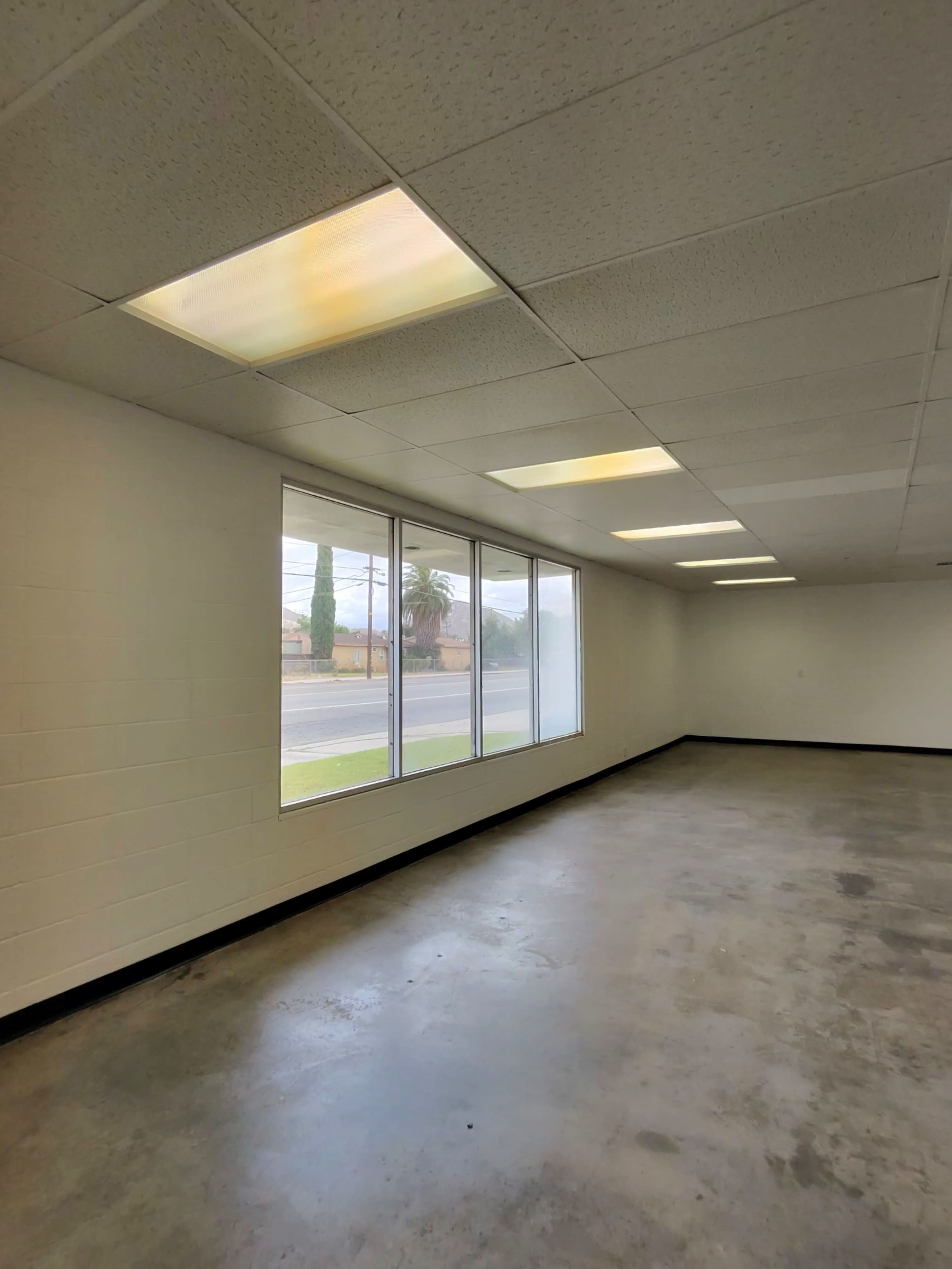 The image shows a vacant, well-lit room with a concrete floor and large windows along one wall.