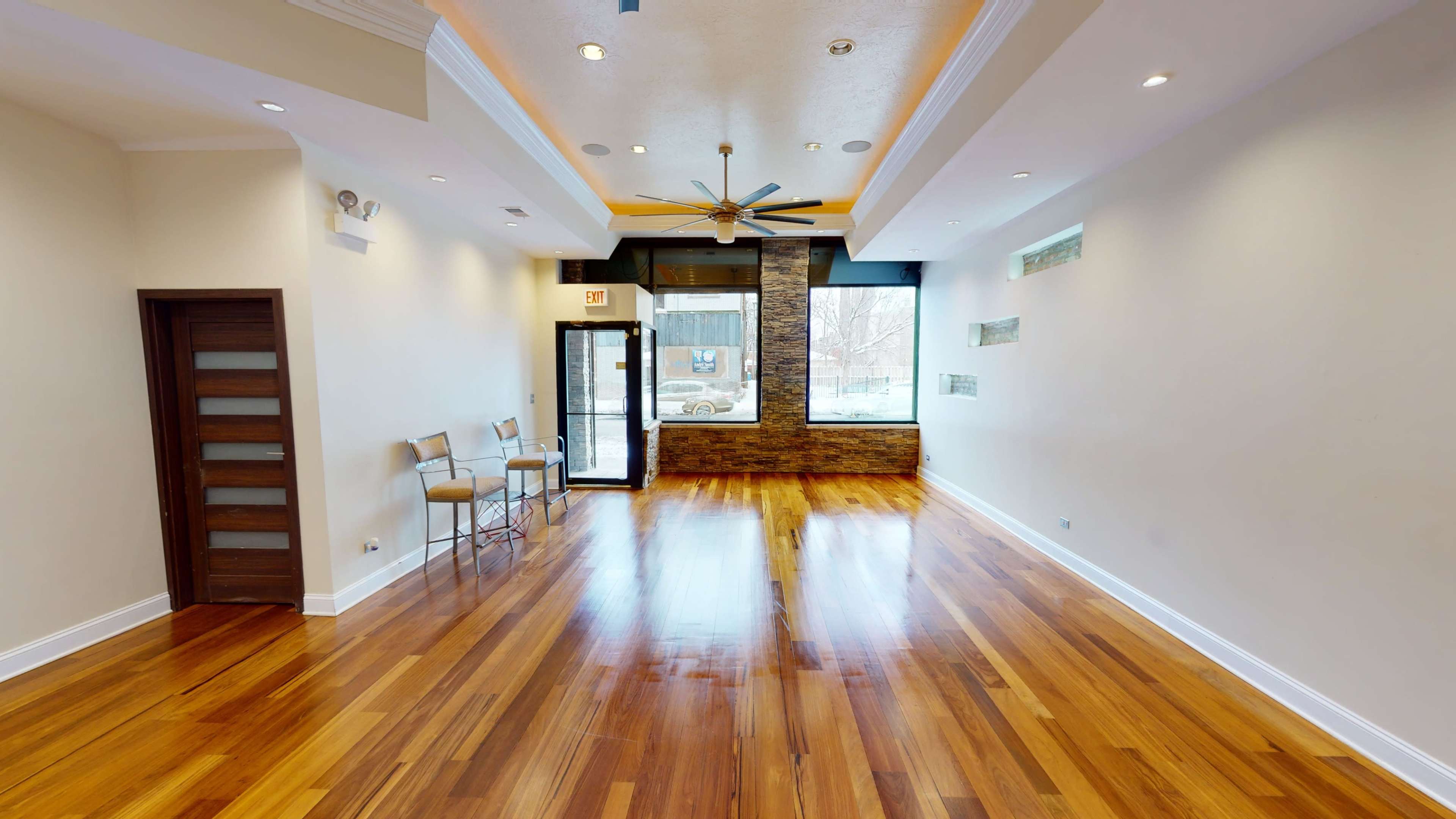 An empty interior space with polished wooden floors, large windows, and a ceiling fan.