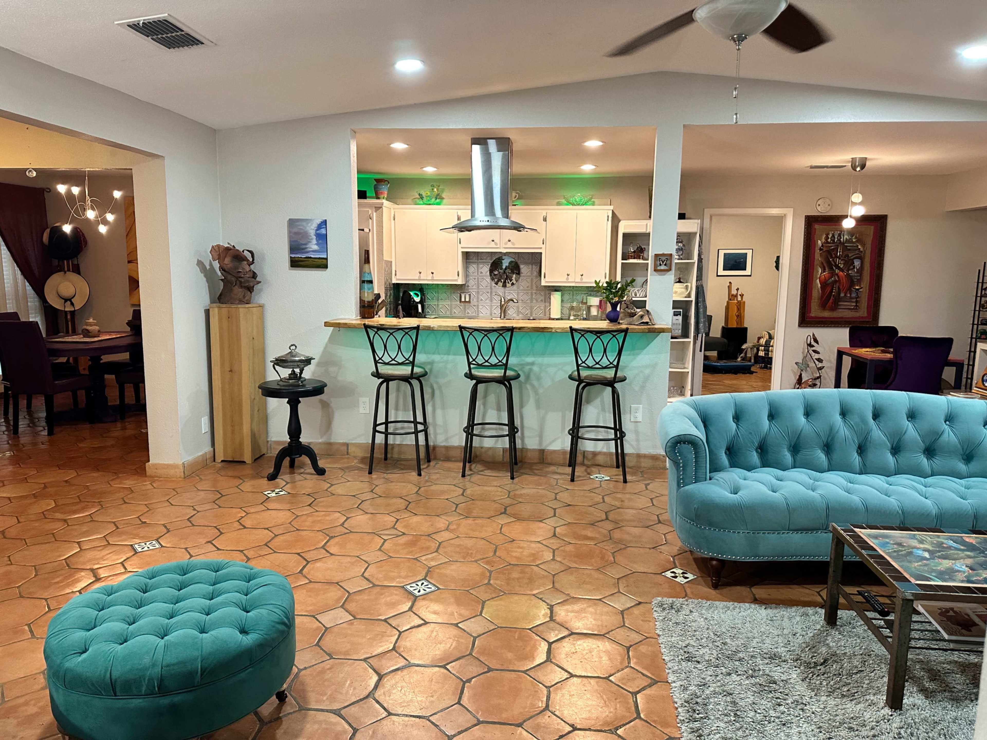 A living area with a turquoise sofa and ottoman, a tile floor, and a kitchen bar with three black stools.