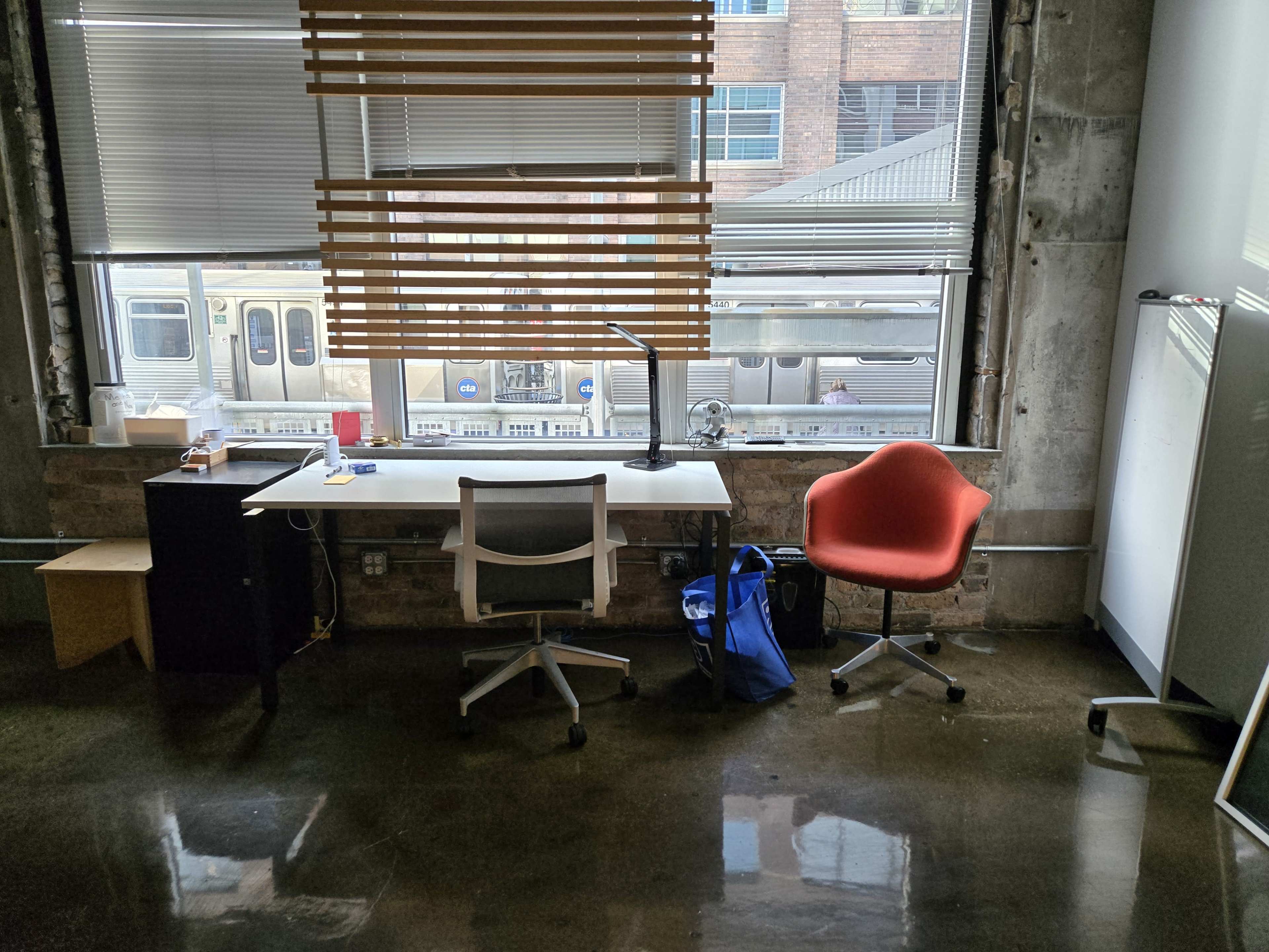 The image shows a workspace with a white desk, an ergonomic chair, a red chair, and a large window overlooking a train passing by outside.