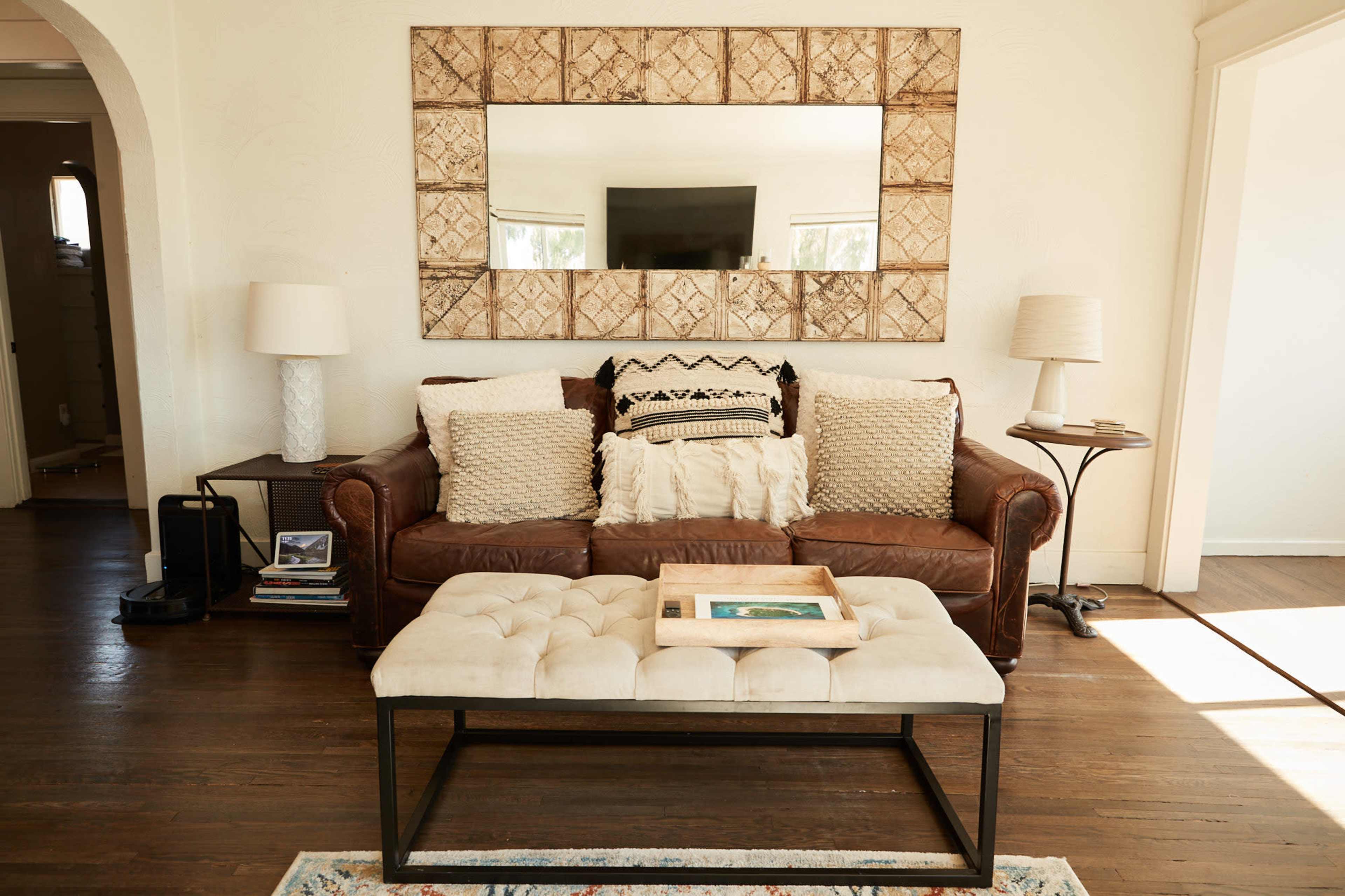 A living room features a brown leather sofa with decorative pillows and a tufted coffee table, set against a large mirror and wooden flooring.