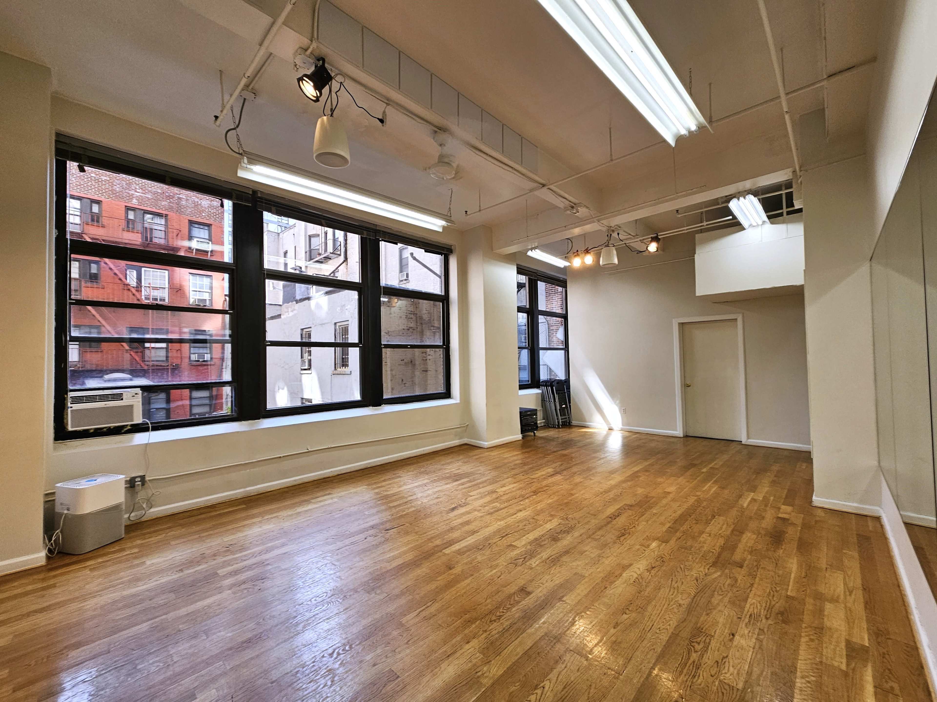 A spacious, empty room with large windows, wooden flooring, and a wall-mounted mirror.