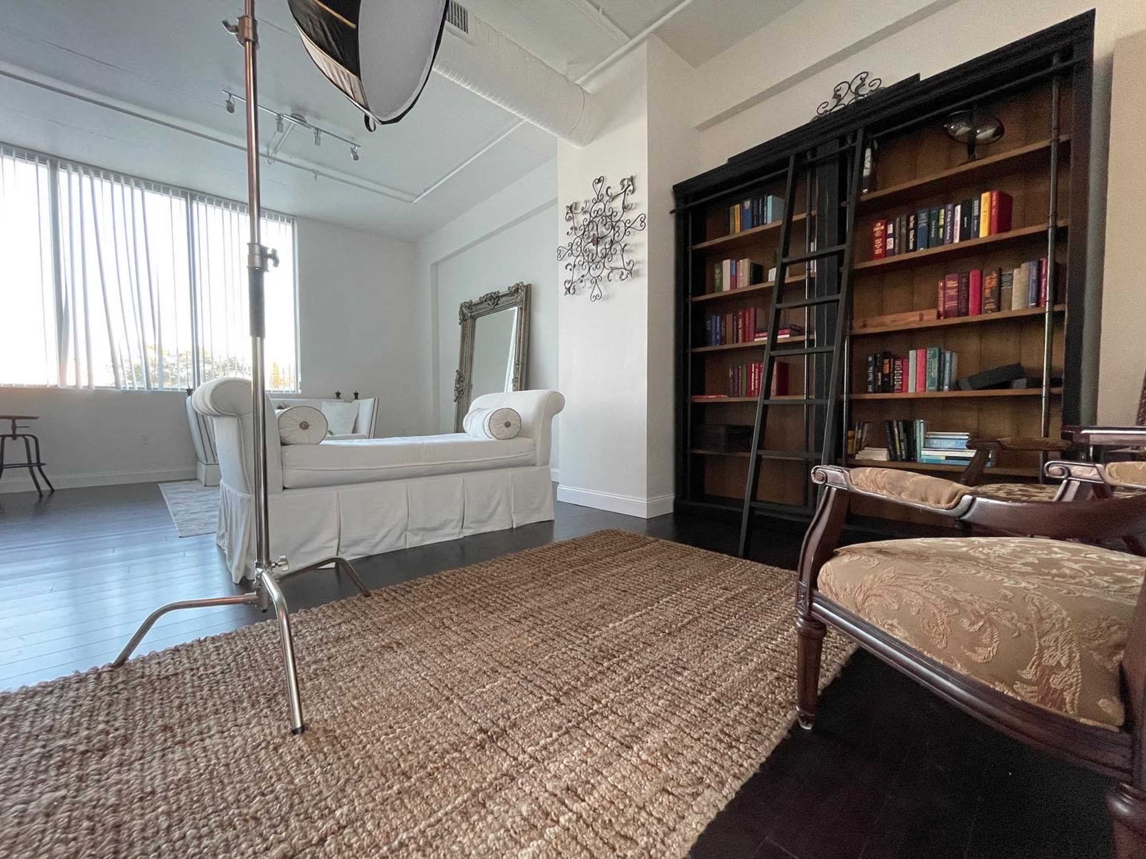 The image shows a spacious room with large windows, featuring a white sofa, a bookshelf filled with books, and a rug on a dark wooden floor.