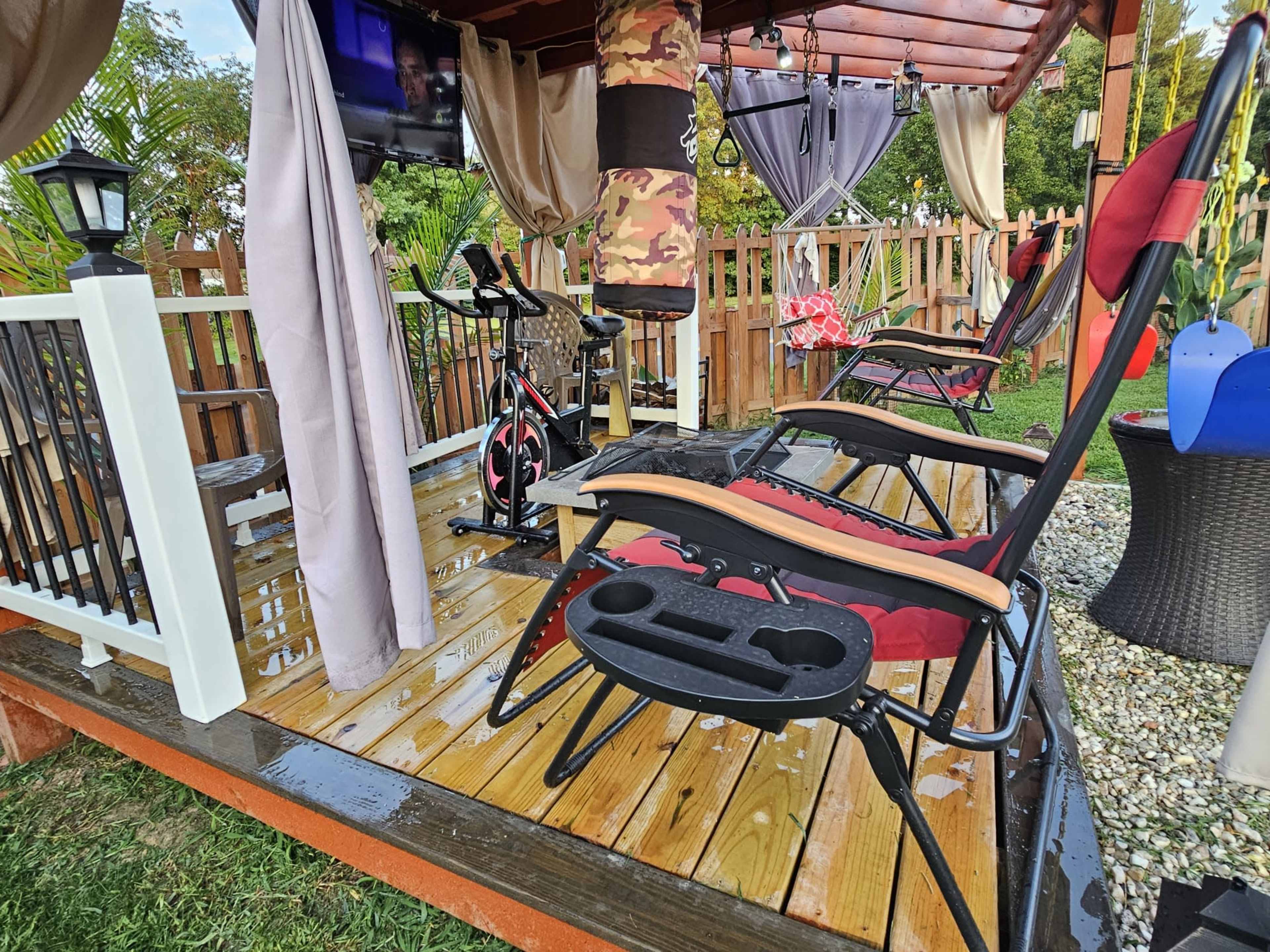 A sheltered outdoor space features reclining chairs and an indoor exercise bike on a wooden deck, with a television mounted nearby.