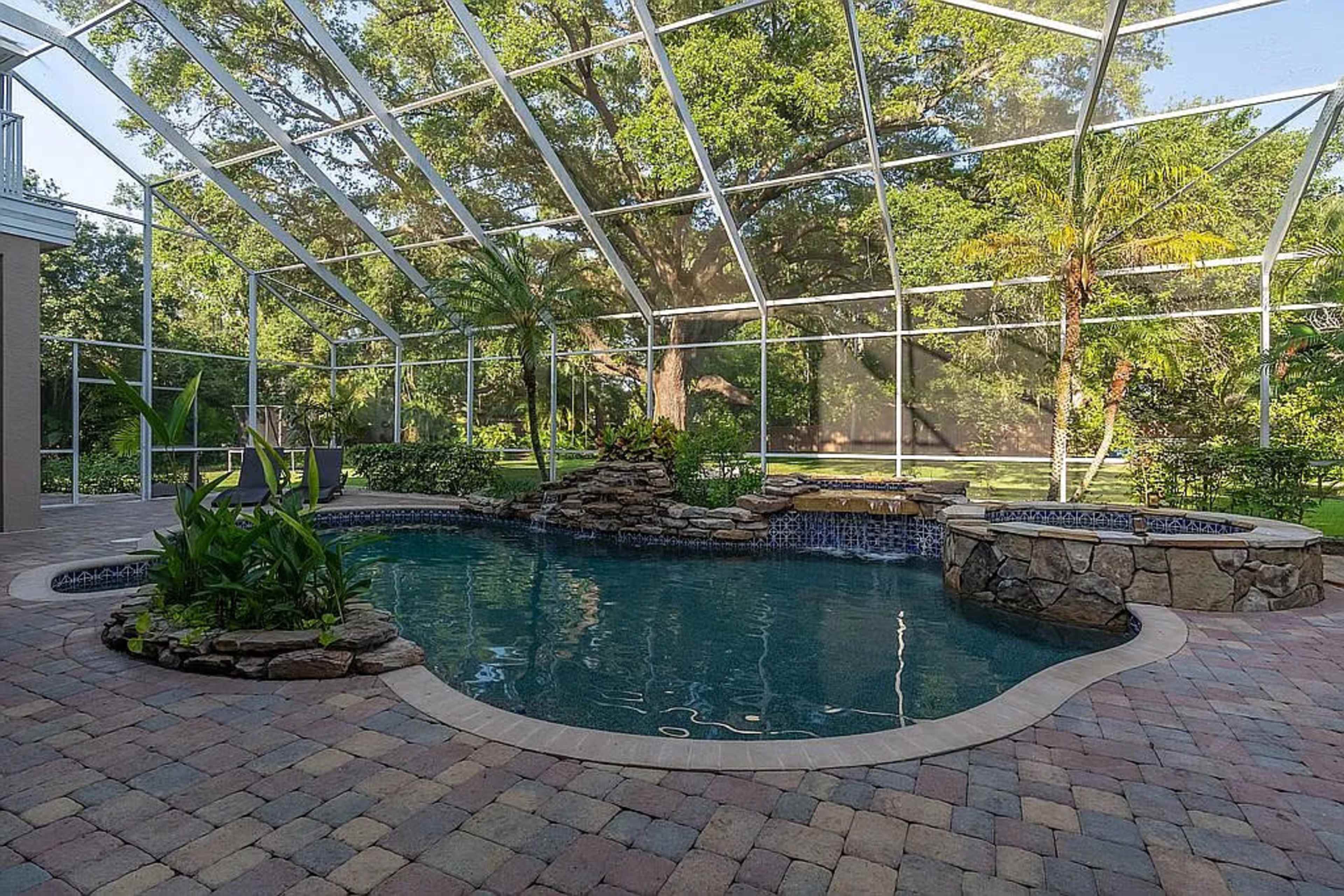 The image shows a rectangular swimming pool surrounded by a stone patio and a screened enclosure, with landscaped greenery in the background.
