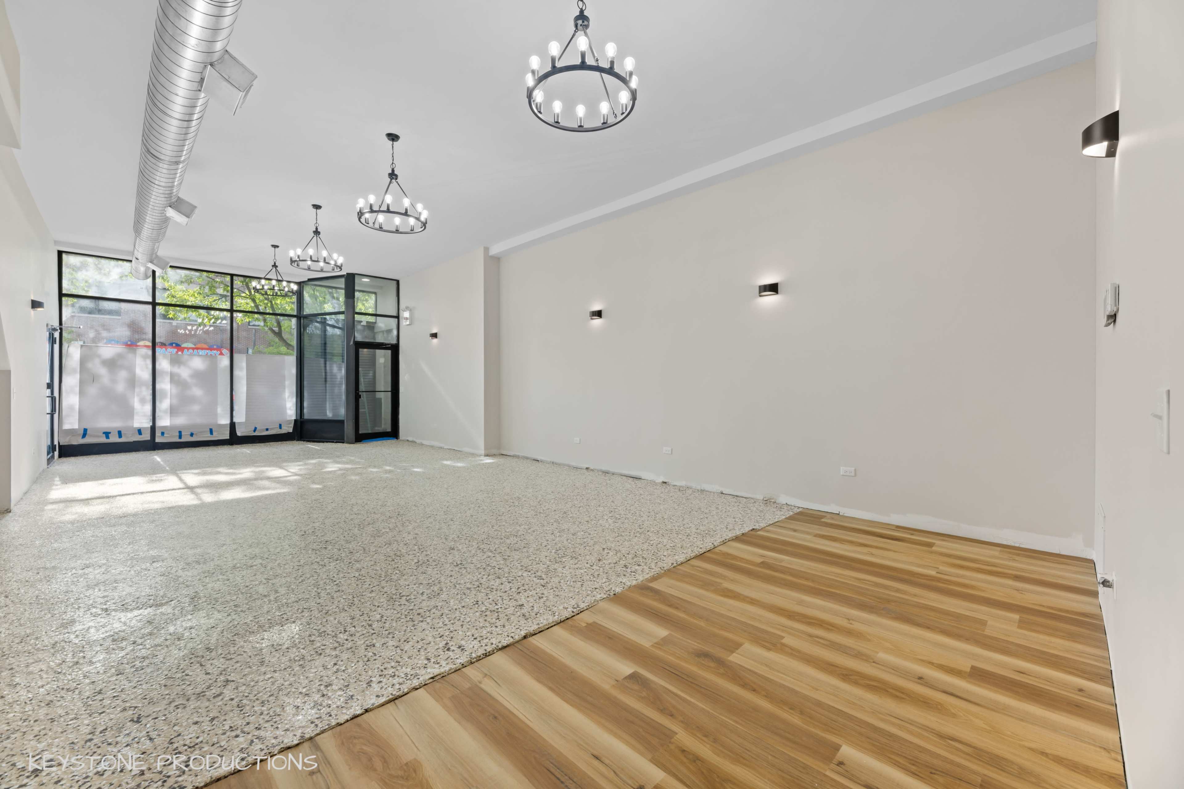 The image shows a spacious commercial room with a mix of polished concrete and wooden flooring, illuminated by several chandeliers and wall sconces.