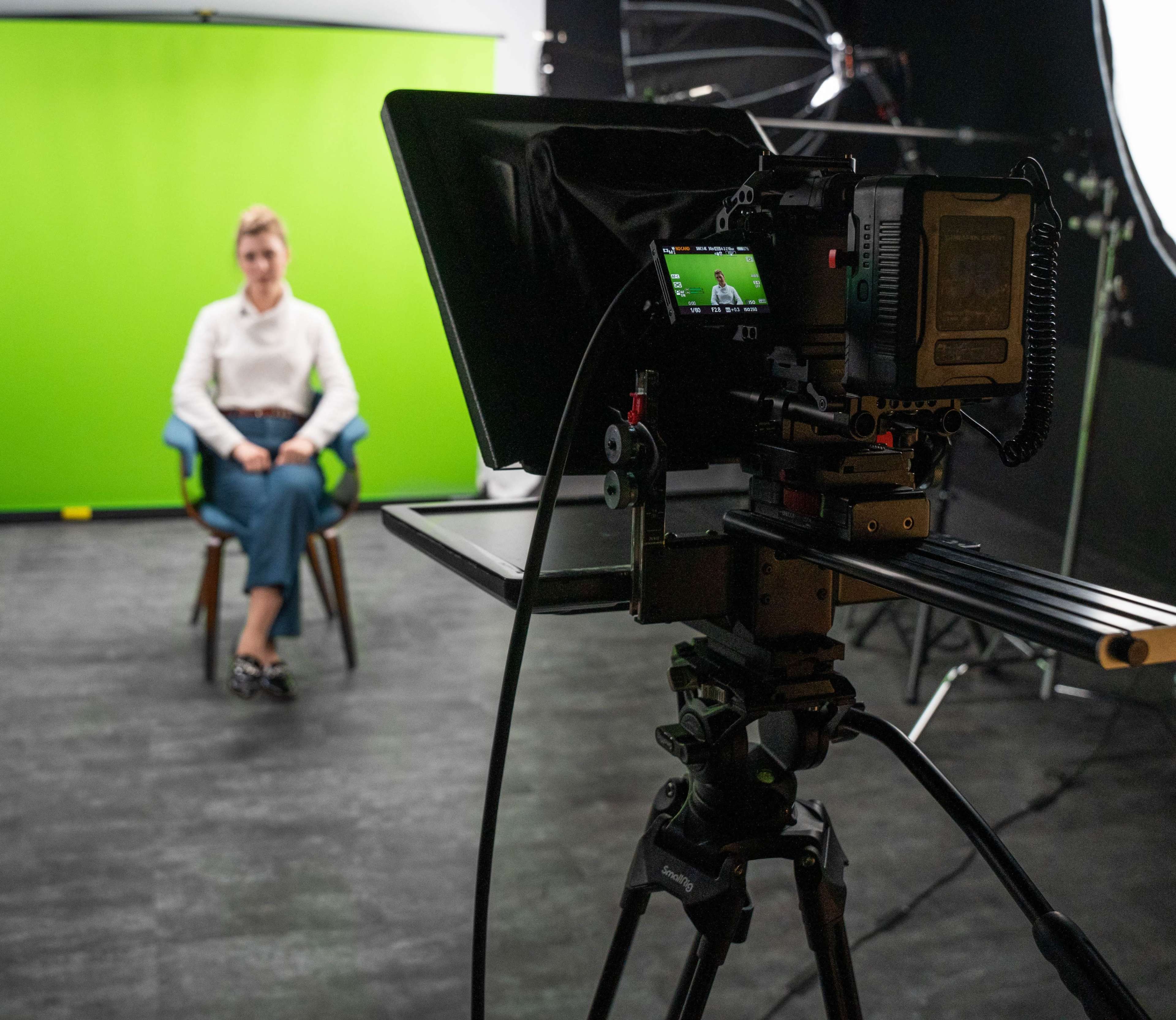 A camera is set up on a tripod facing a person seated in front of a green screen in a studio setting.