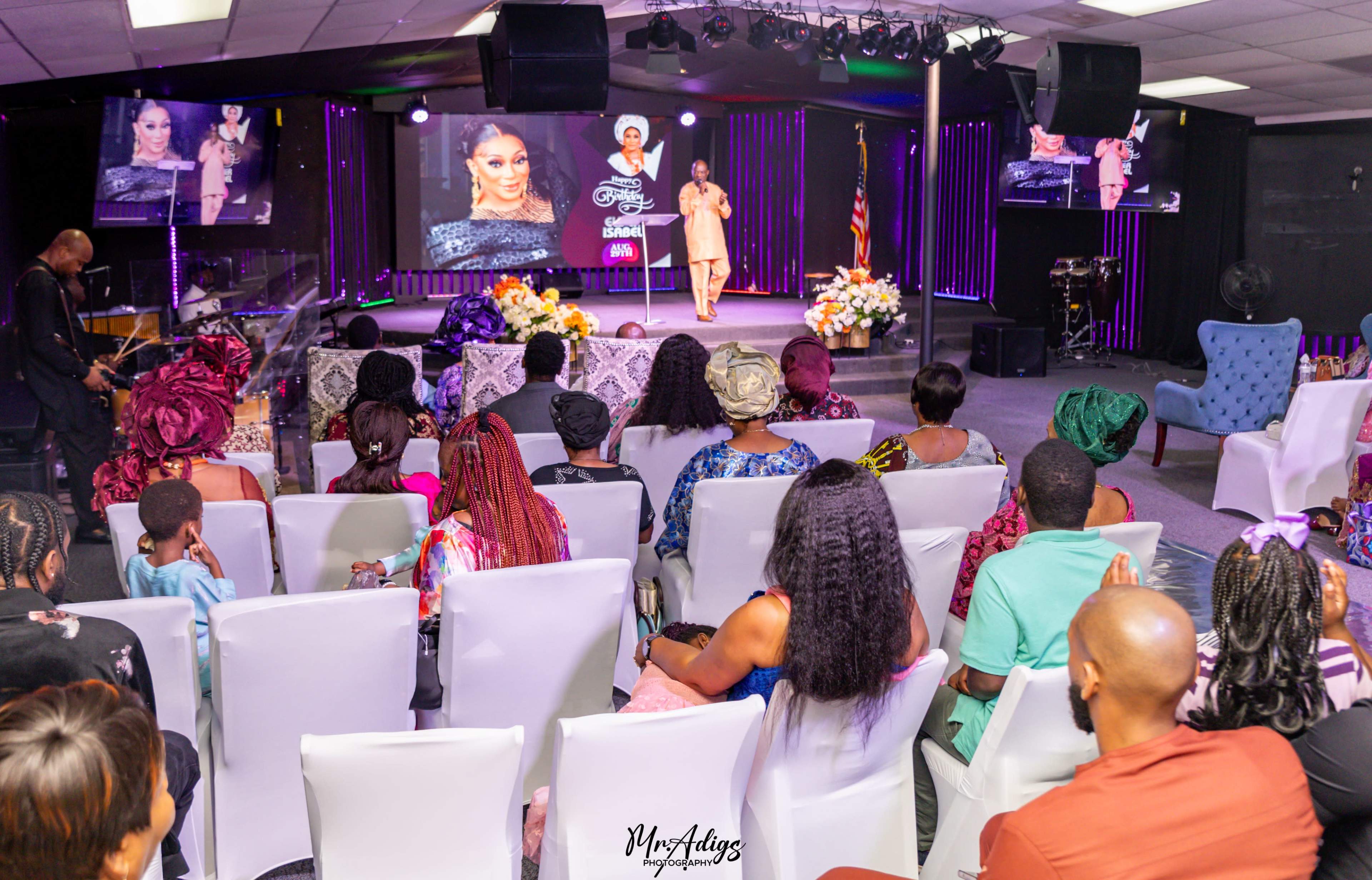 A crowd gathers in a well-lit hall, watching a speaker on stage, with vibrant decorations and large screens displaying images.