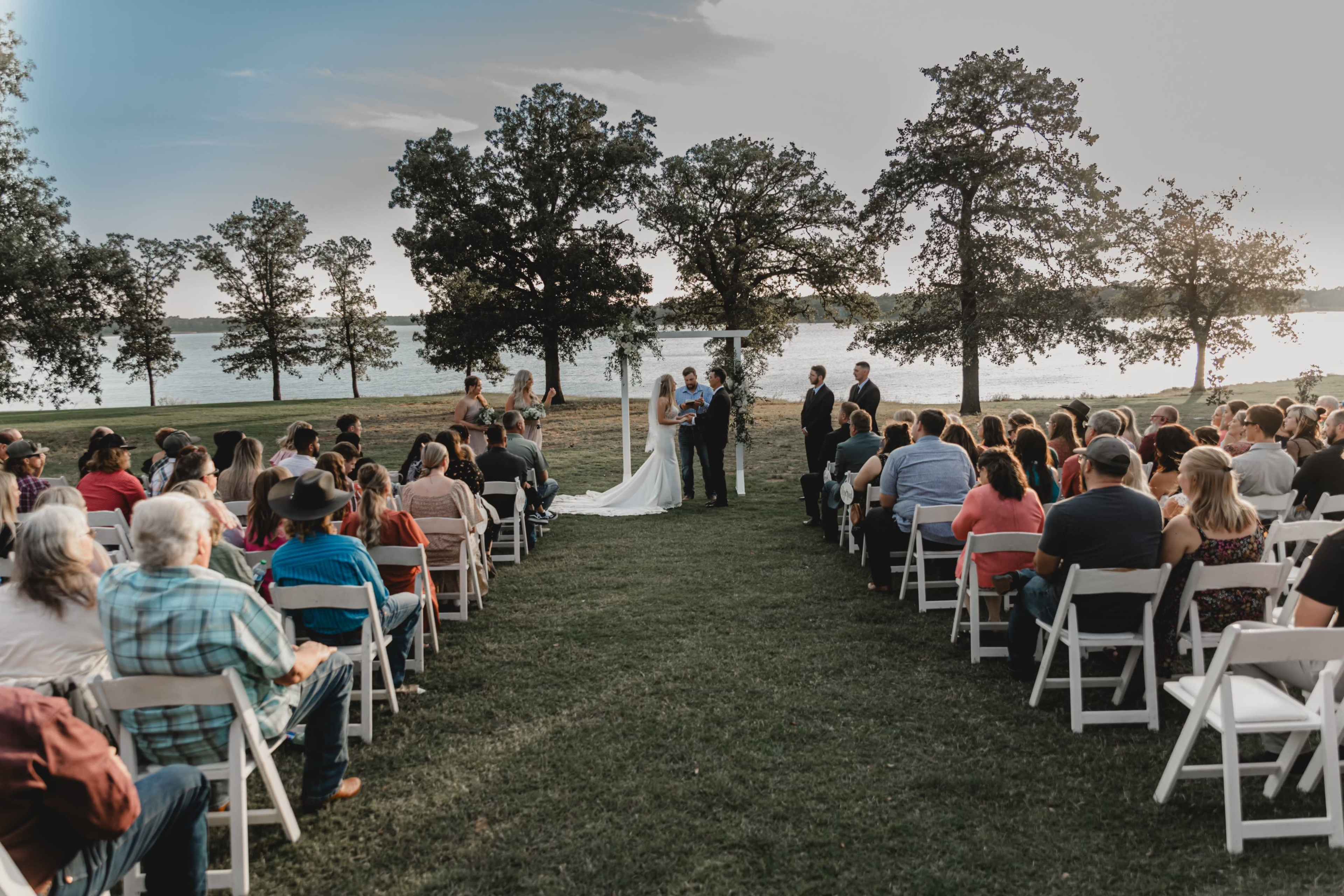 A couple stands under an arch exchanging vows in front of an audience seated on white chairs beside a body of water surrounded by trees.