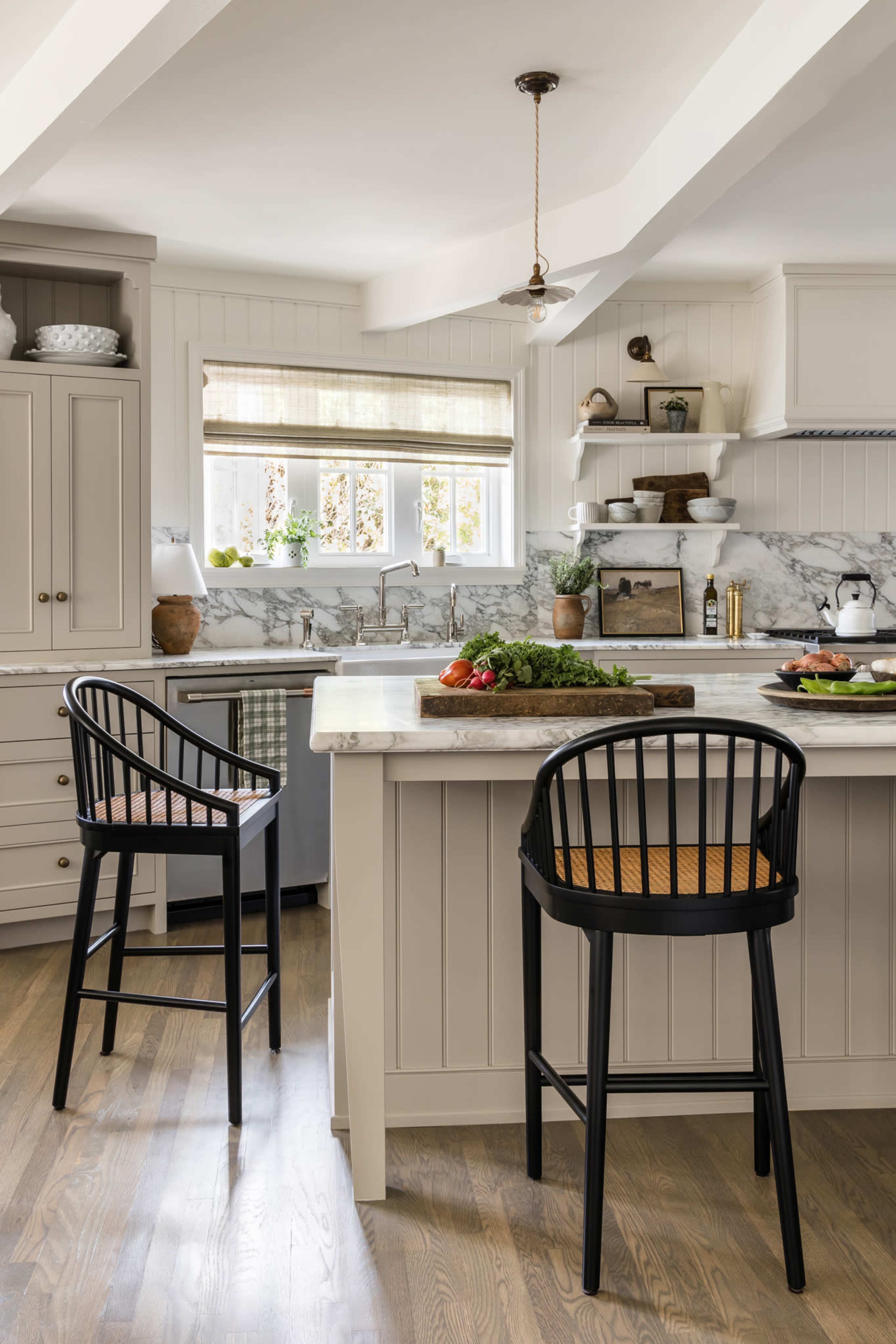 A modern kitchen features a marble countertop with two barstools, white cabinetry, and natural light streaming through a window.