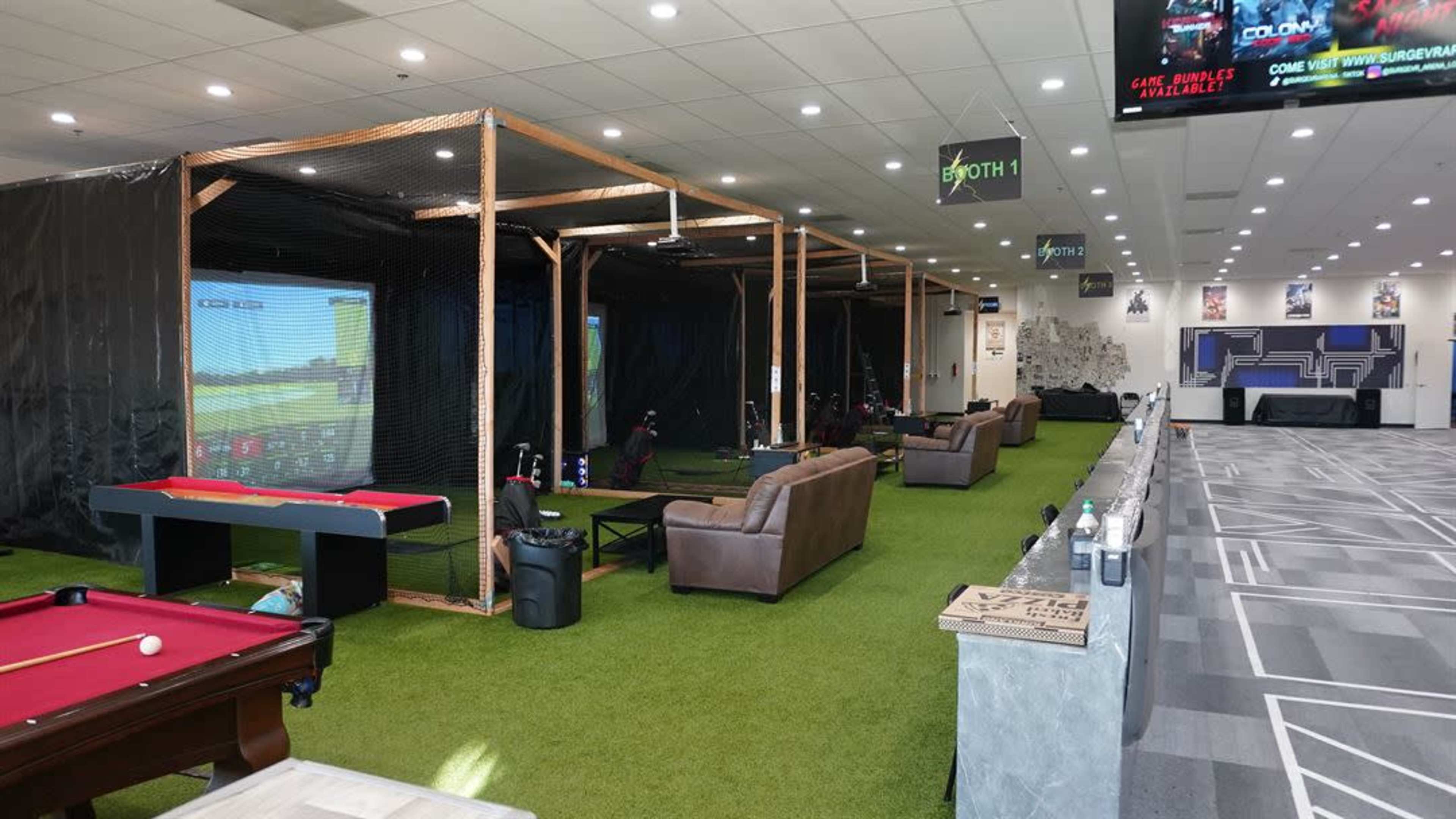 The image shows an indoor recreational space featuring golf simulators, a billiards table, and seating areas on artificial grass flooring.