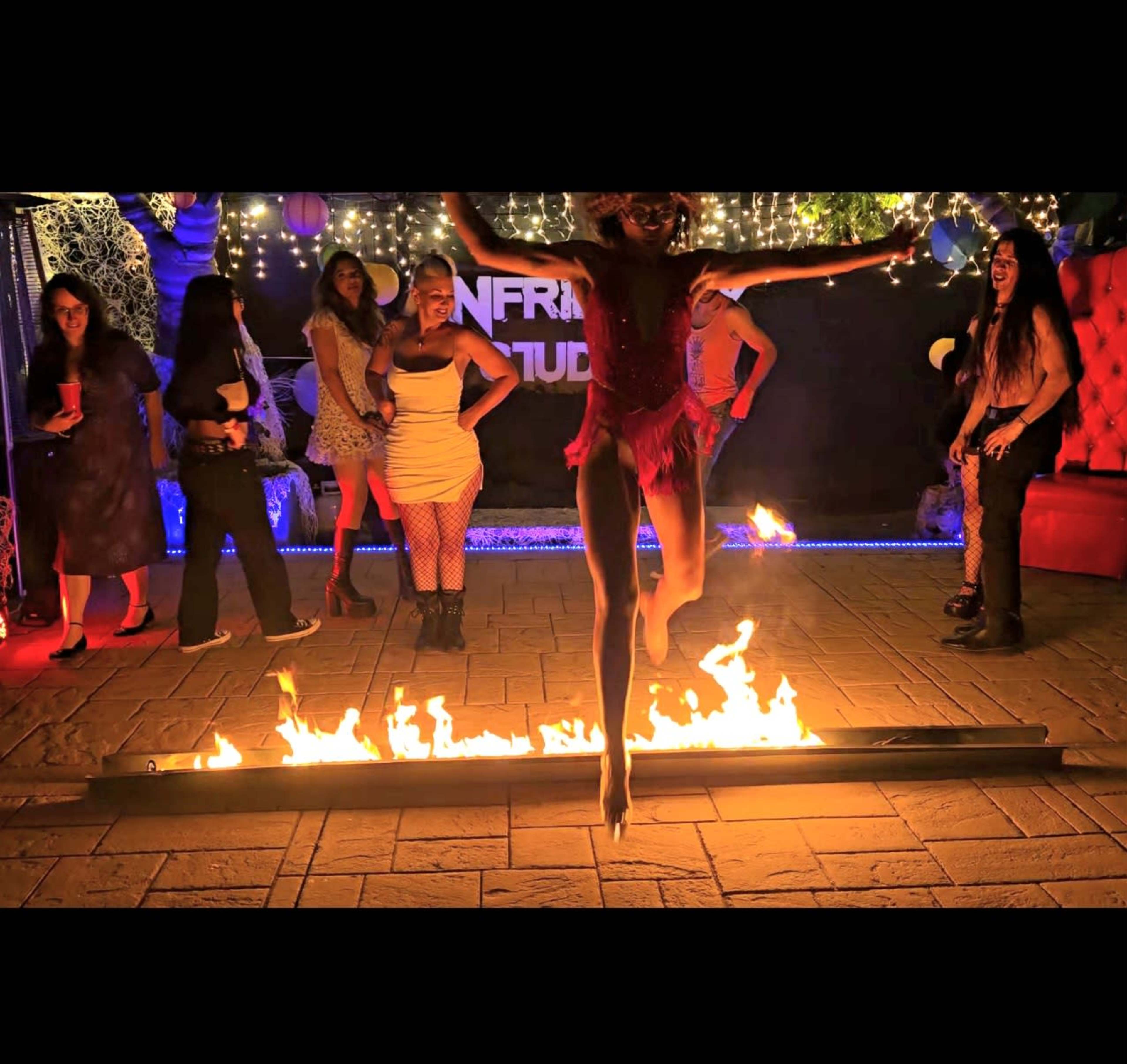 A performer in a pink outfit jumps over a line of fire while several spectators watch in a brightly lit party setting.