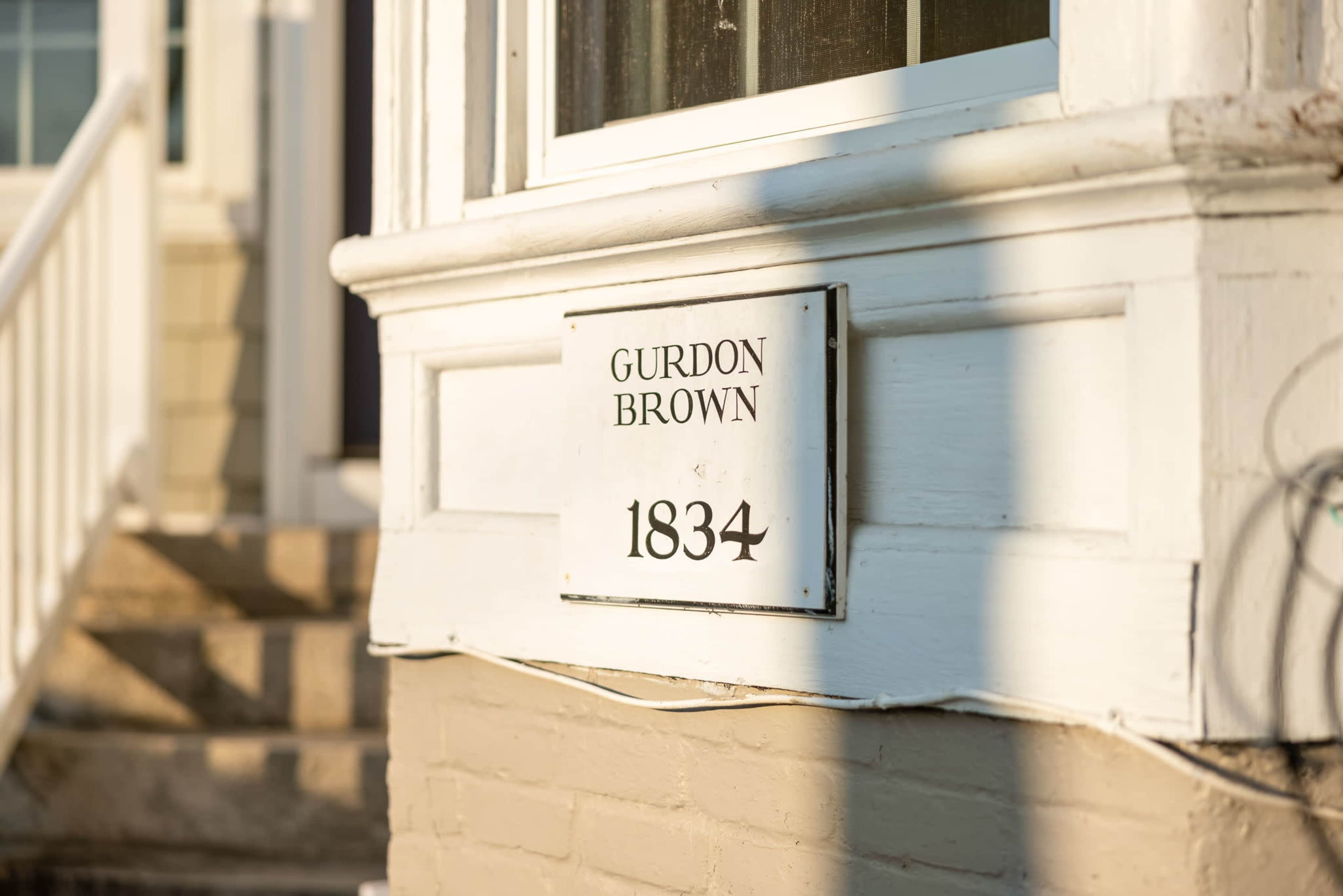 A house address sign that reads "Gurdon Brown 1834," mounted on a white exterior wall near a staircase.