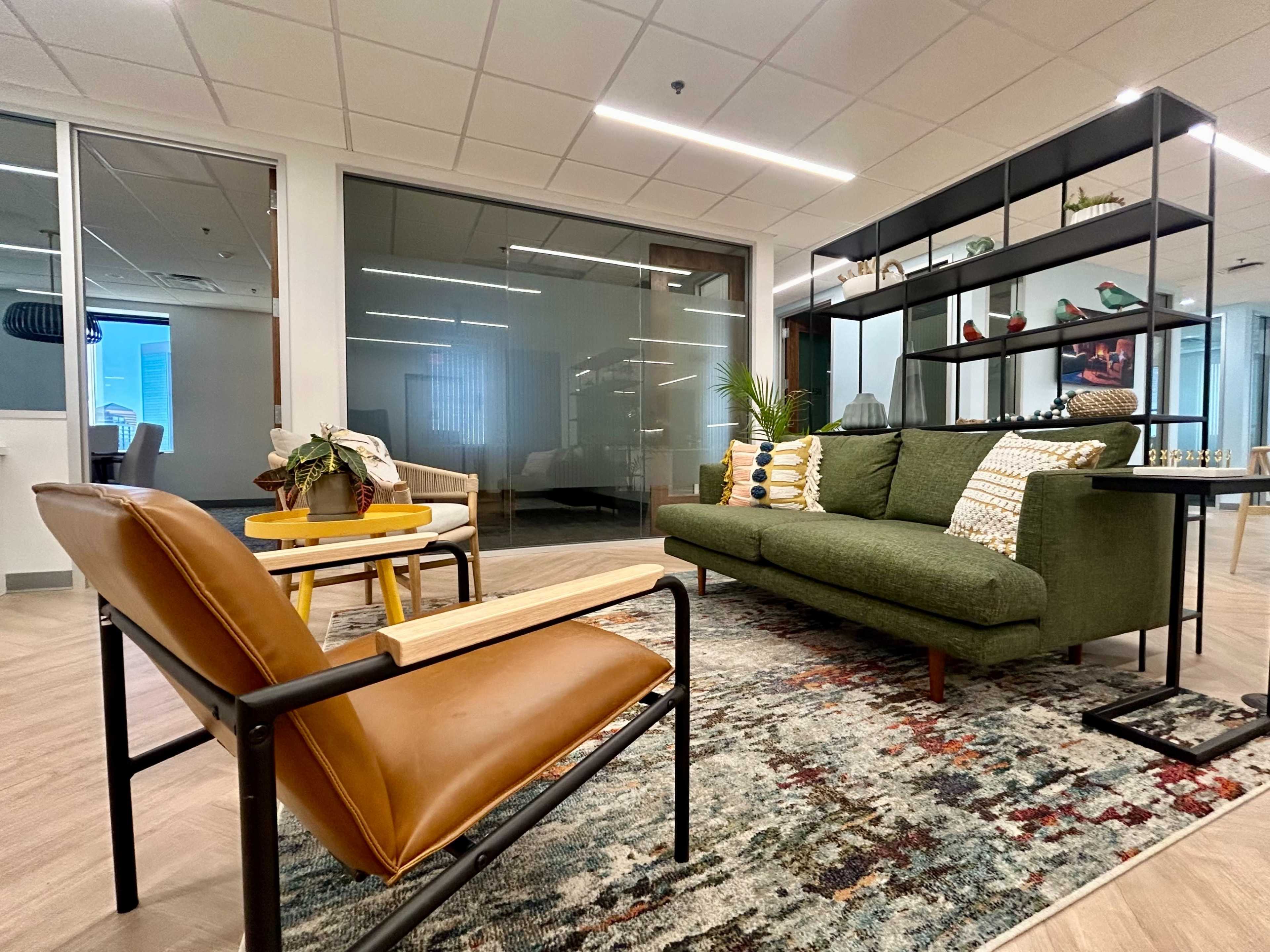 The image shows a modern office lounge area featuring a green sofa, two armchairs, a wooden side table, and shelves with decorative items.