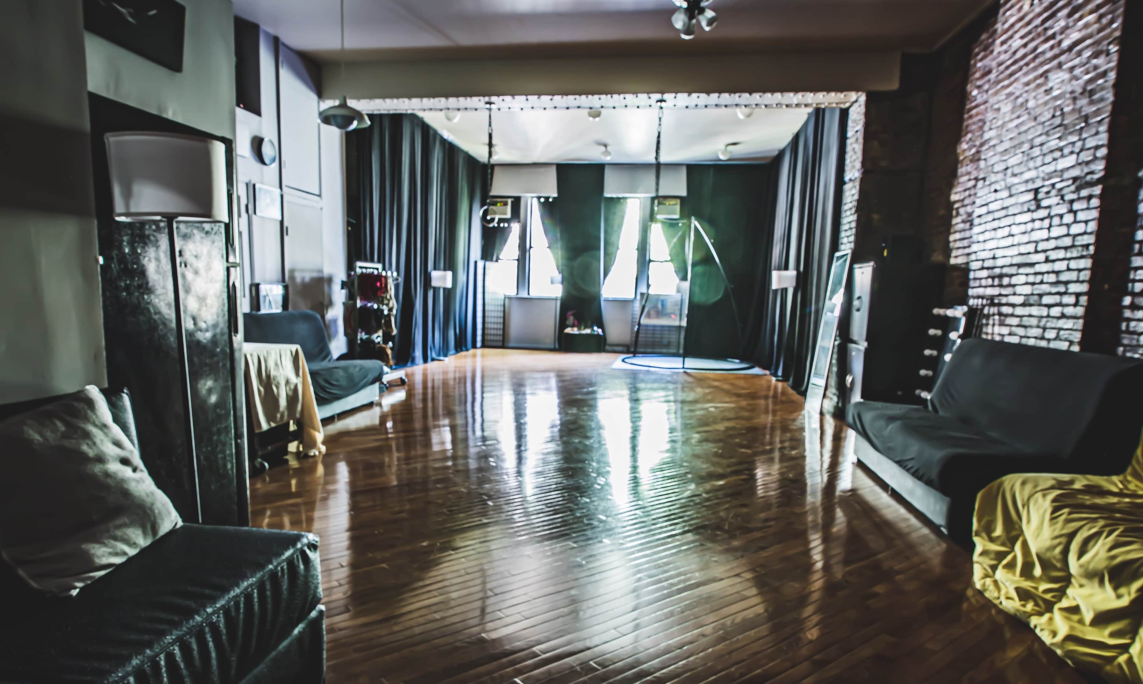 The image shows a spacious room with polished wooden floors, large windows, and dark curtains, furnished with couches and various equipment along the walls.