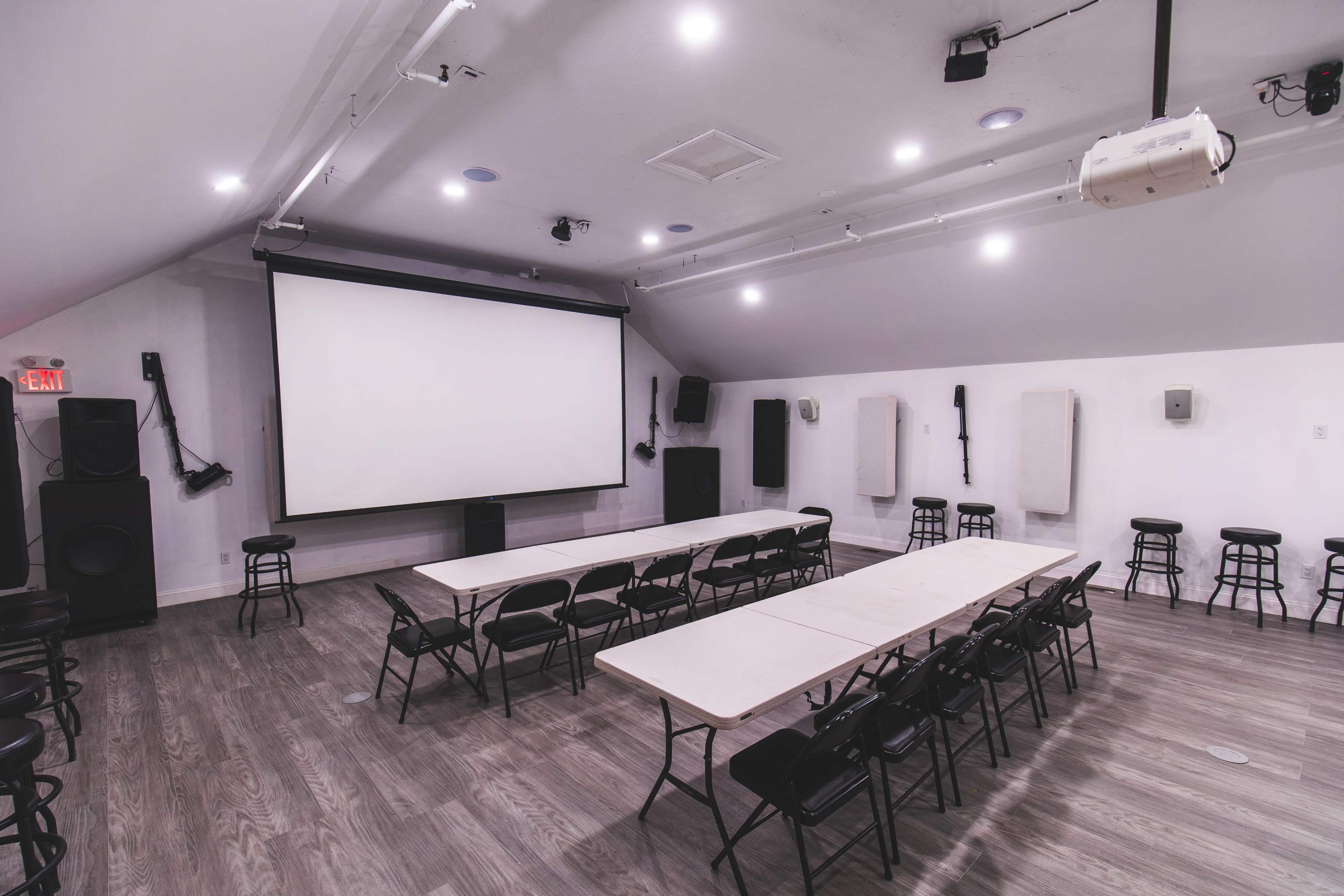The image shows an empty multi-purpose room with a large screen at the front, sound equipment on the sides, and rows of chairs arranged around several tables.
