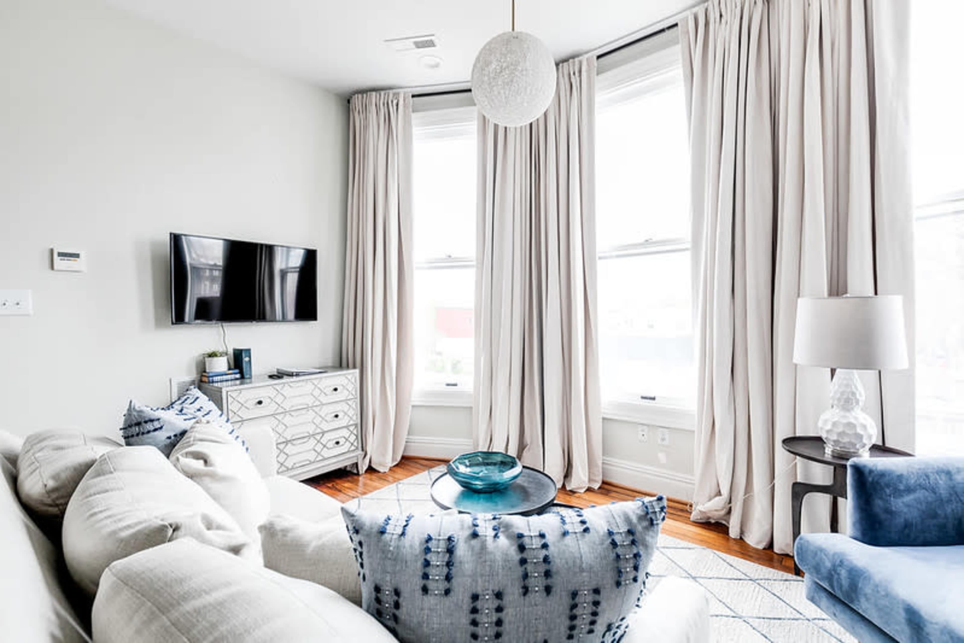 A modern living room features a sectional sofa with patterned pillows, a round coffee table, a TV mounted on the wall, and large windows draped with light-colored curtains.