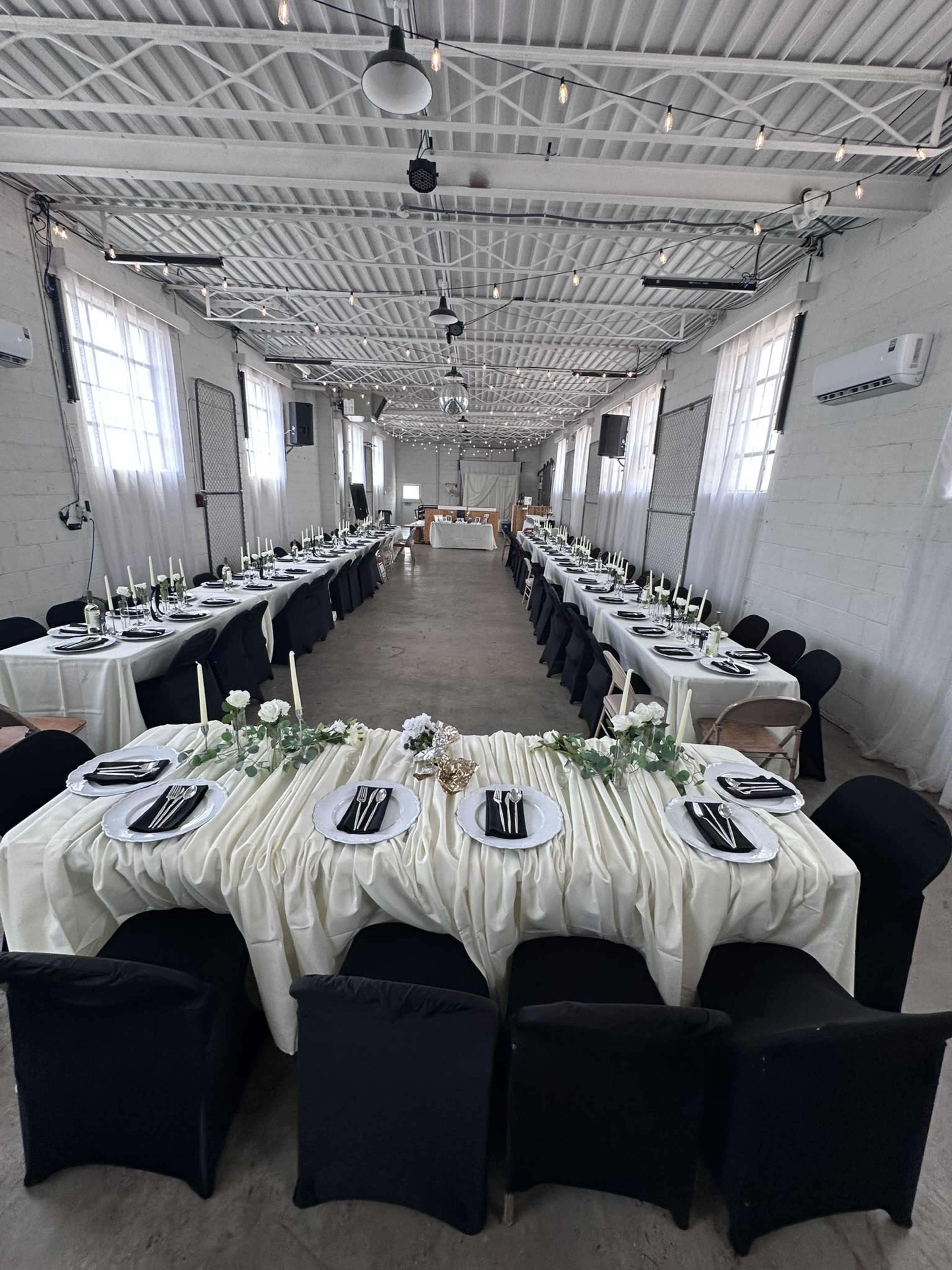 A spacious indoor venue features two long tables elegantly set with white tablecloths and black chairs, adorned with candles and floral centerpieces.
