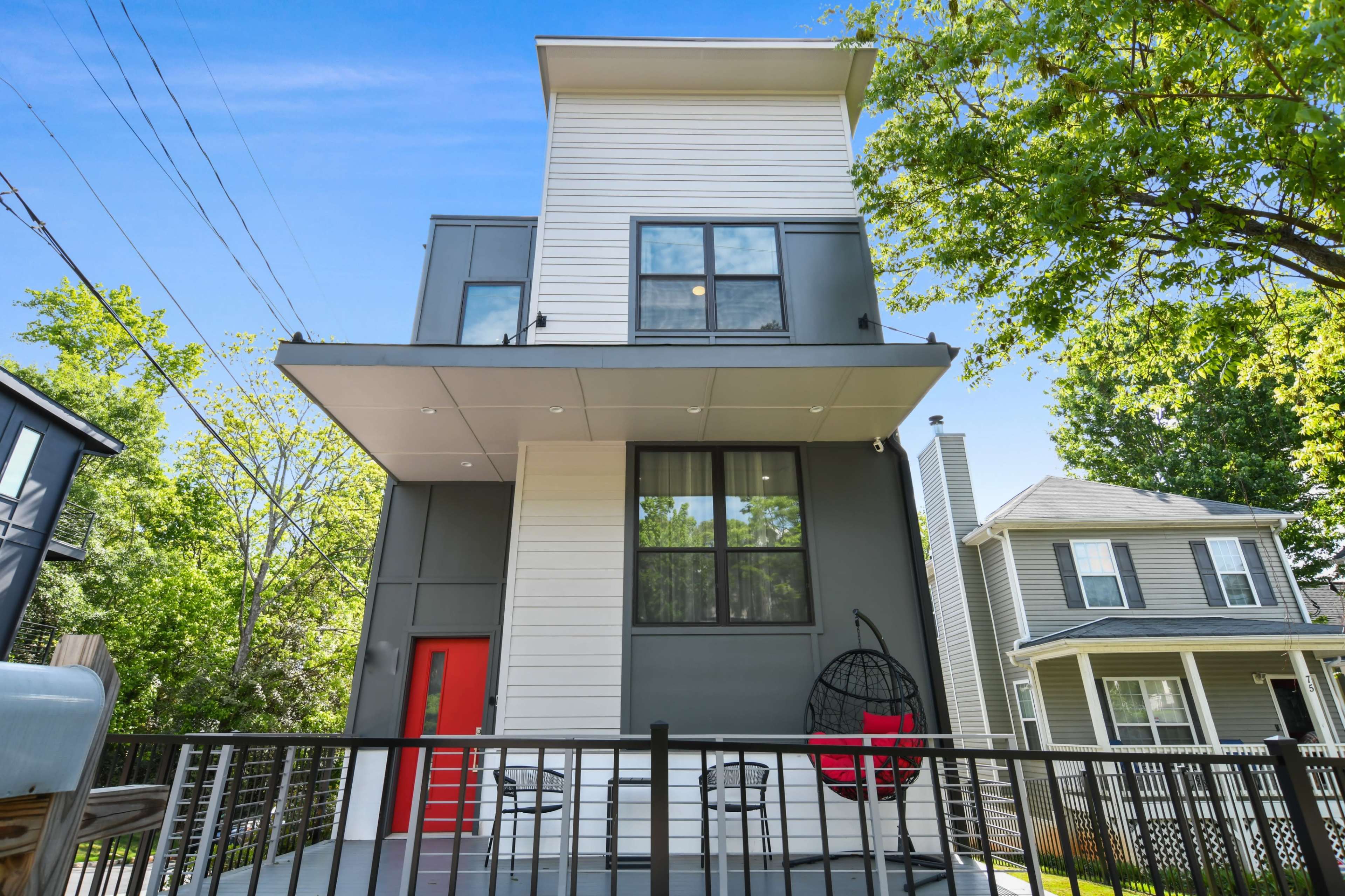 A modern two-story house with a flat roof, featuring a red front door and a balcony, surrounded by trees.