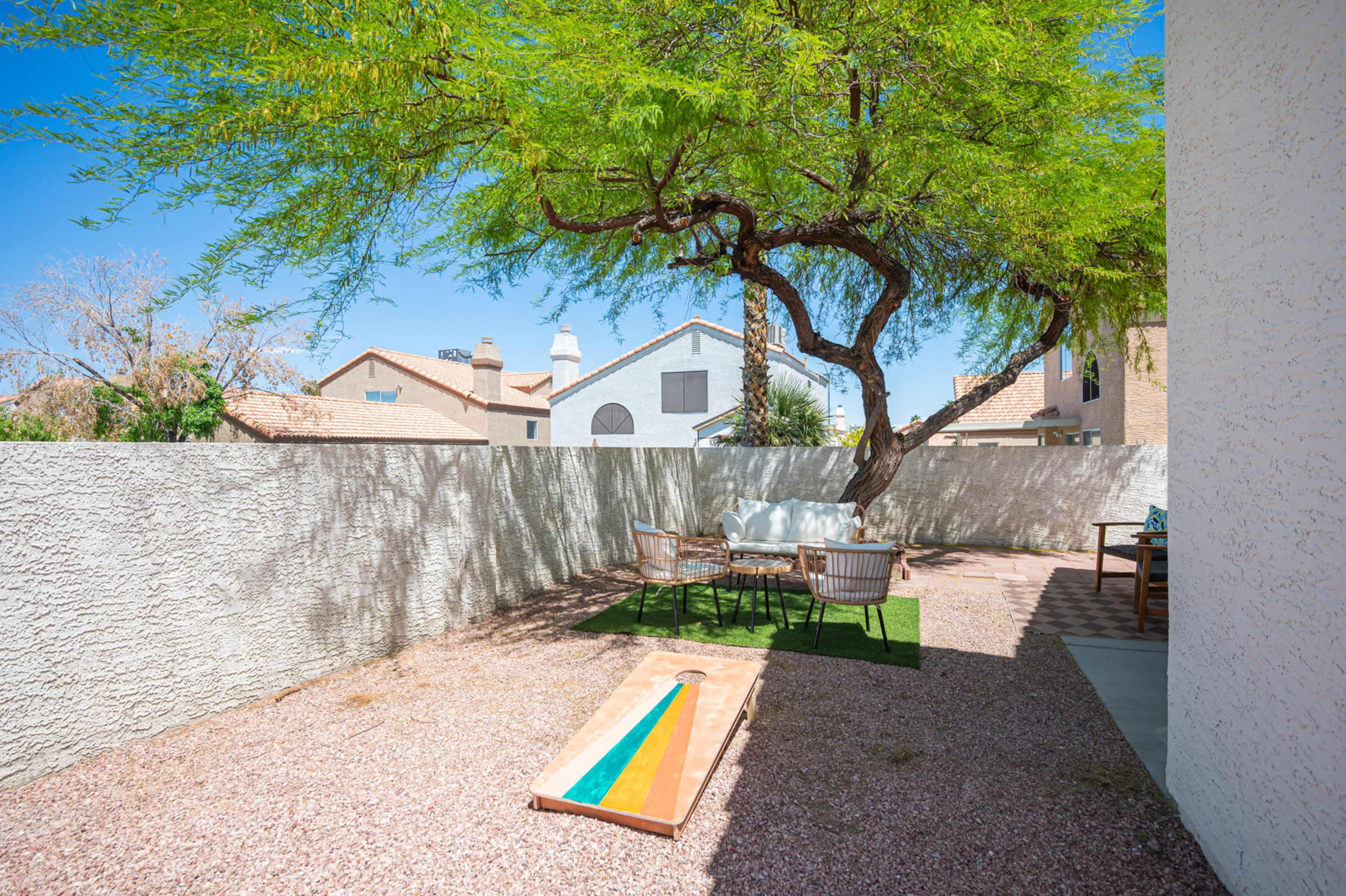 A backyard scene features a gravel area with a large tree, a table and chairs set on grass, and a cornhole game in the foreground.