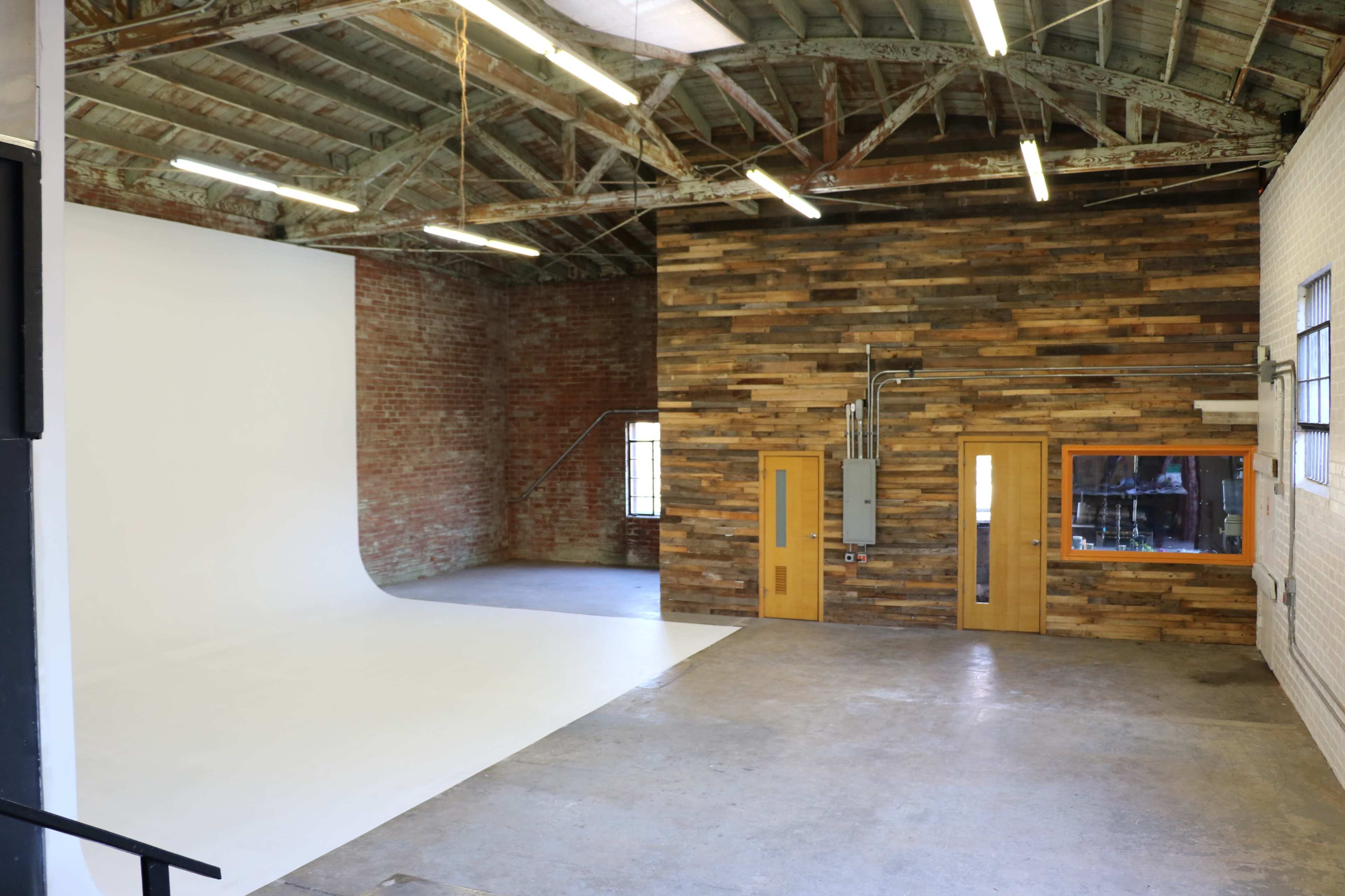 The image shows a spacious studio interior featuring a white backdrop on one side and a wooden accent wall with windows on the opposite side.