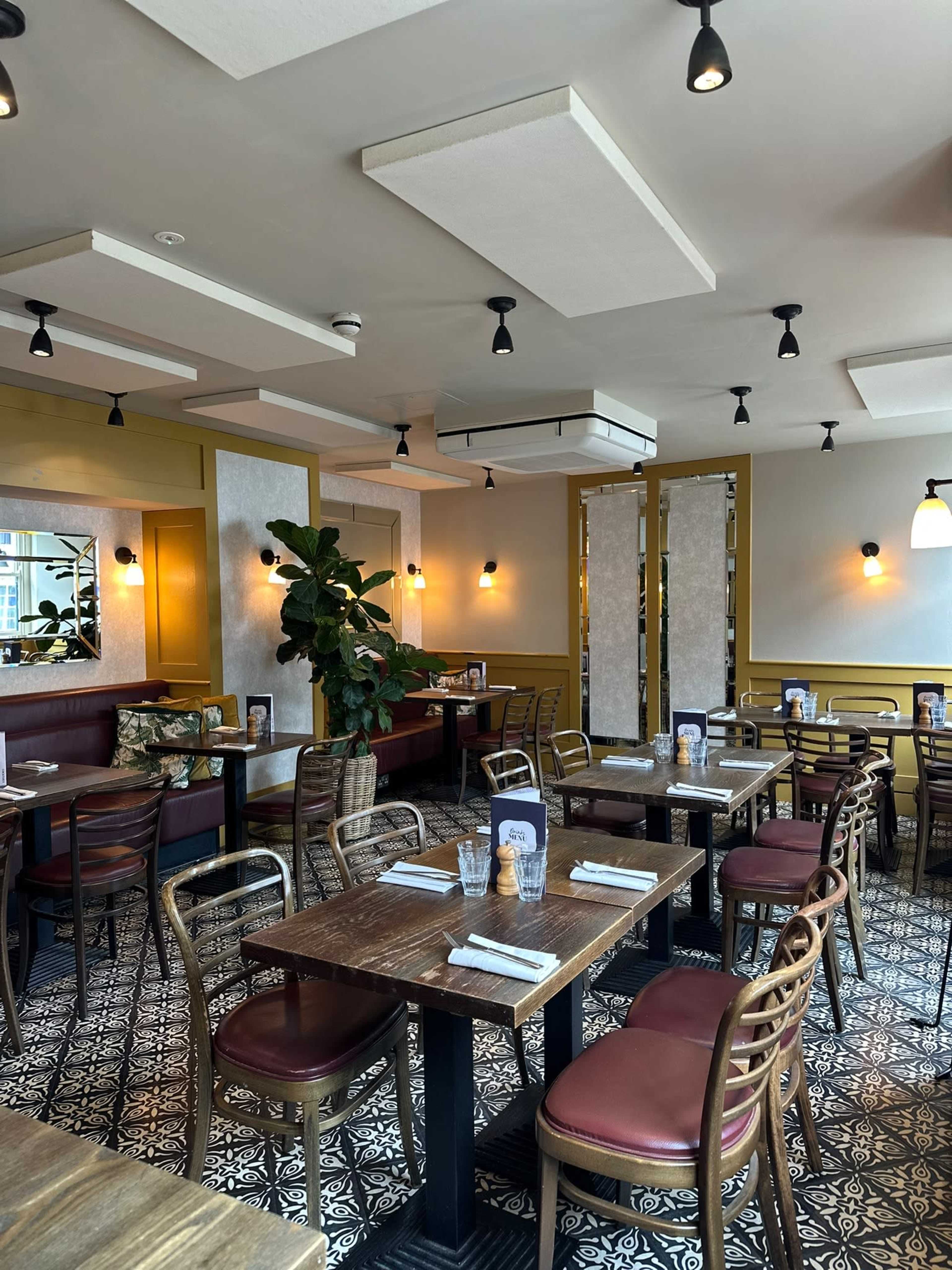 The image shows a modern restaurant interior with wooden tables, red and wooden seating, and patterned tile flooring, illuminated by stylish ceiling lights.