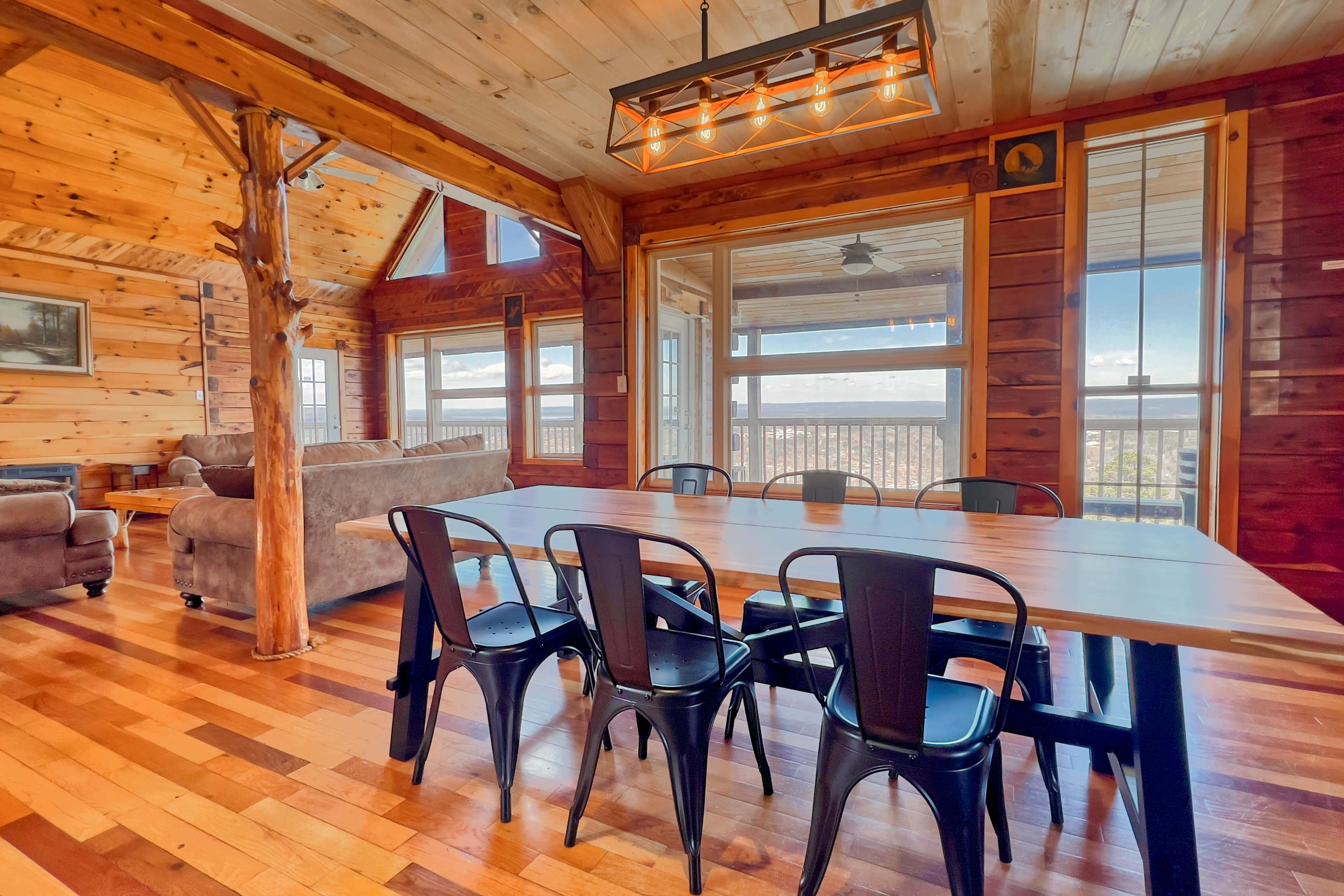 A wooden cabin interior featuring a dining table with black metal chairs, a sitting area with a gray couch, and large windows that provide a view.