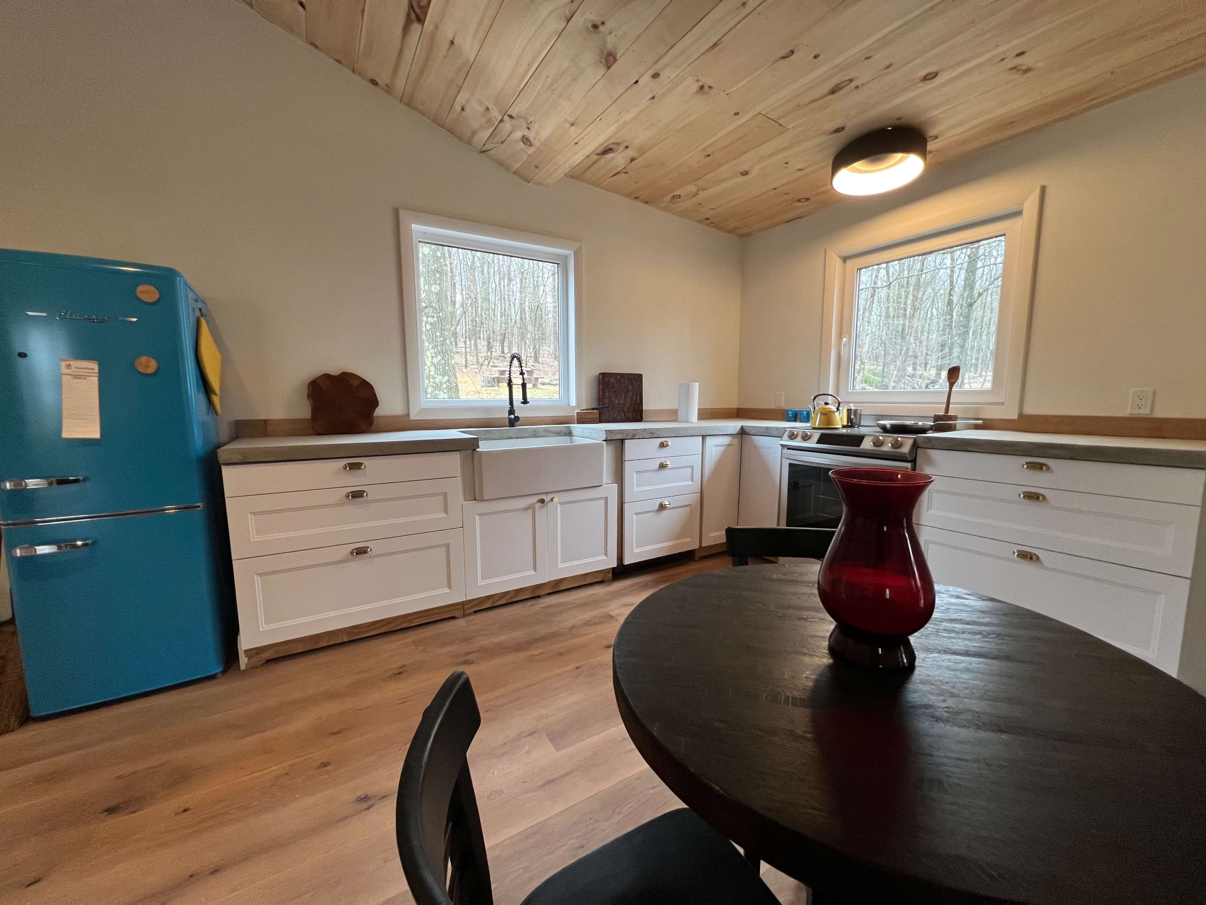 A modern kitchen with white cabinets, a blue retro refrigerator, a round black table, and large windows overlooking a forested area.