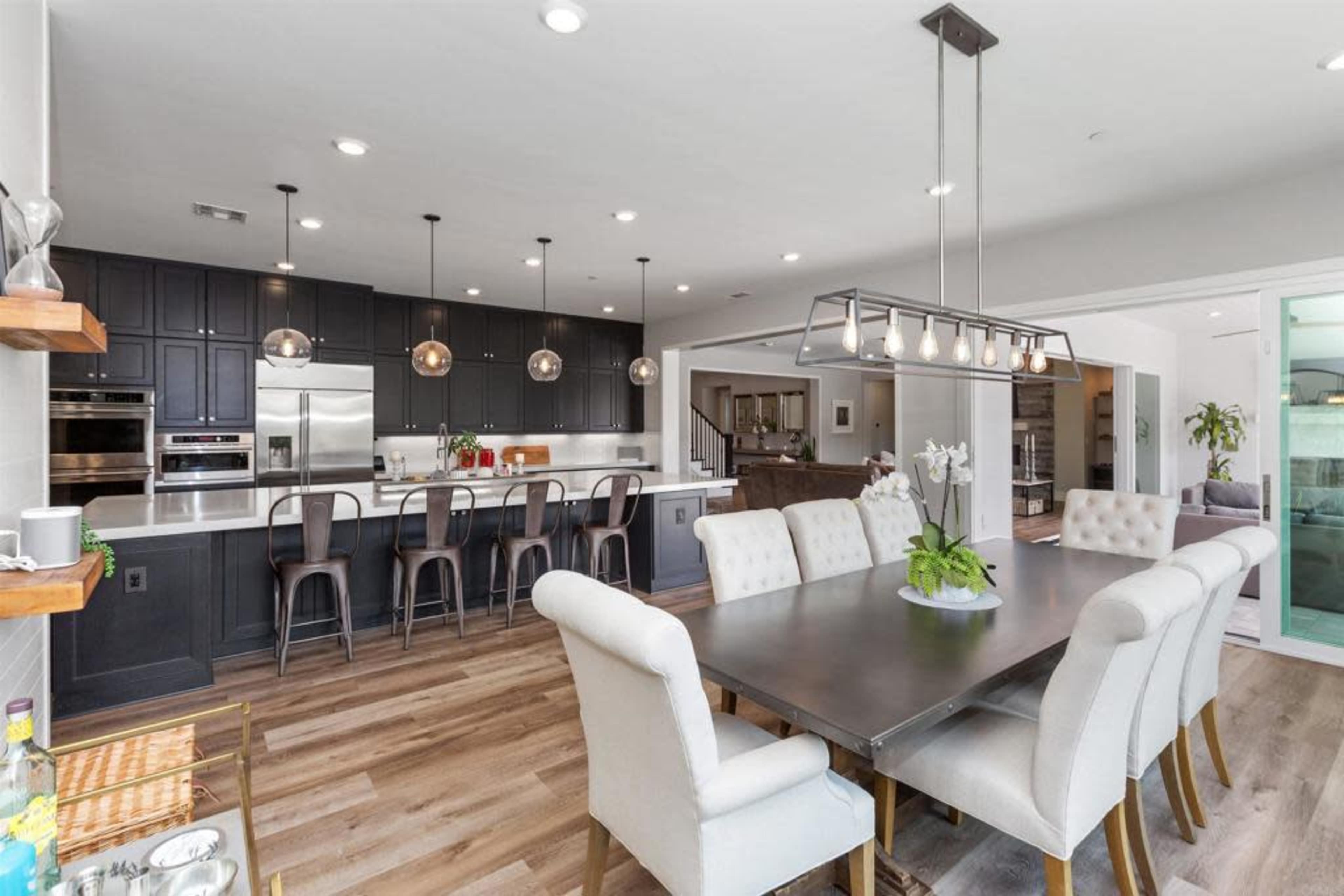 A modern kitchen and dining area features a long table with upholstered chairs, an island with bar stools, and dark cabinetry illuminated by pendant lights.
