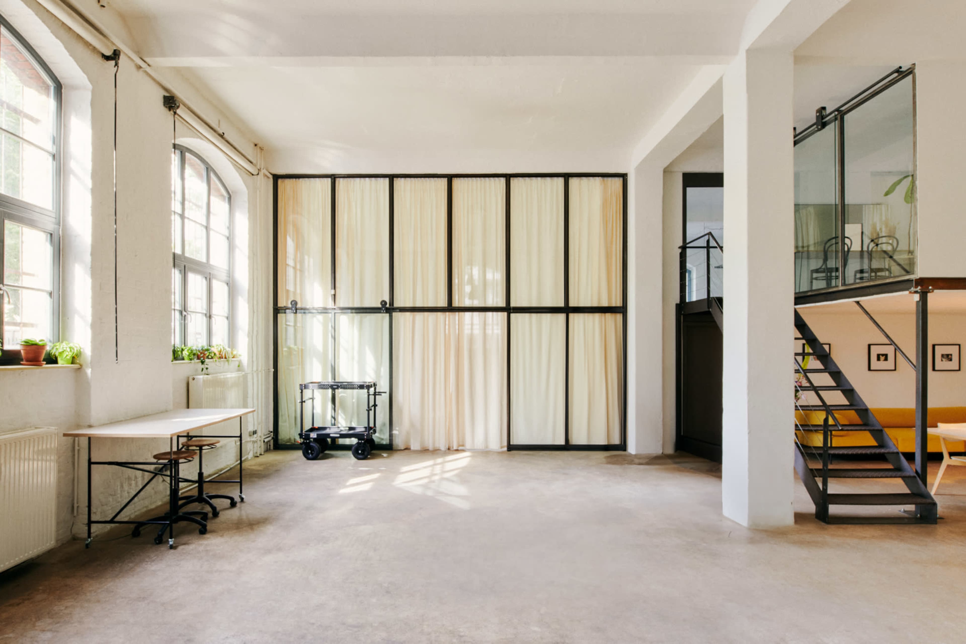 The image shows a spacious, bright interior with large windows, a curtain wall dividing the space, and a staircase leading to an upper level.