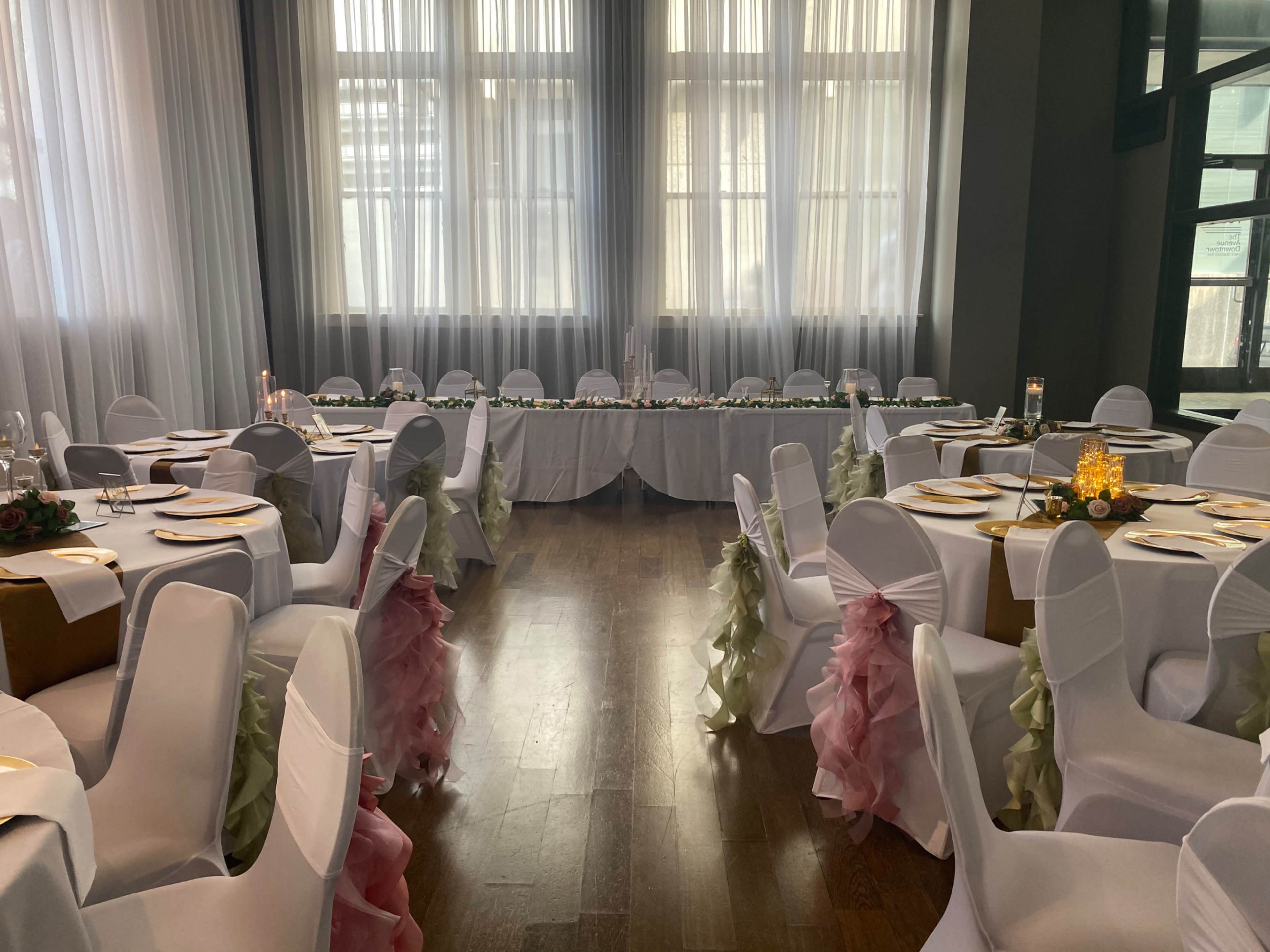 A banquet hall set up for an event, featuring tables adorned with white tablecloths, plates, and chairs draped in fabric with pink and green sashes.