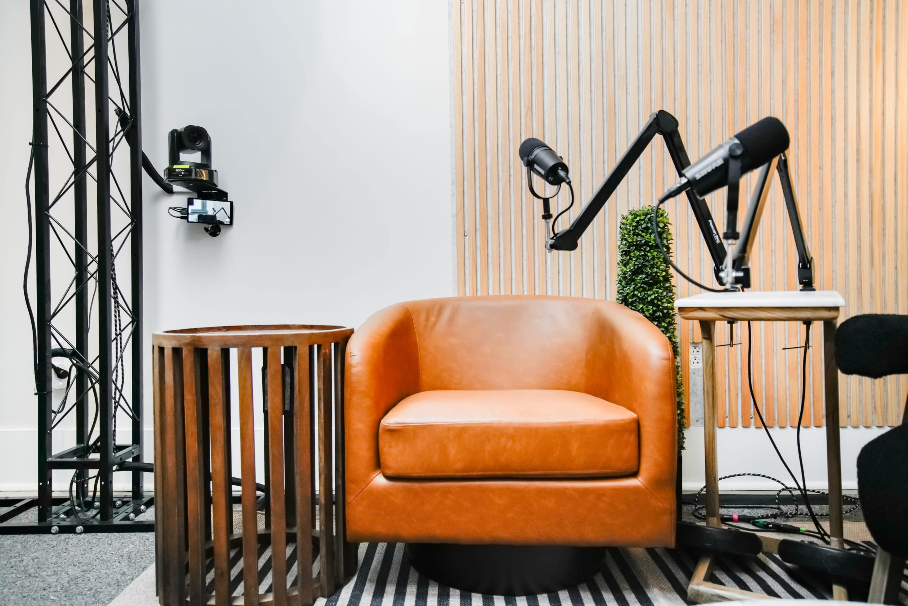 The image features a cozy brown armchair next to a wooden side table and a desk with adjustable microphone stands in a studio setting.