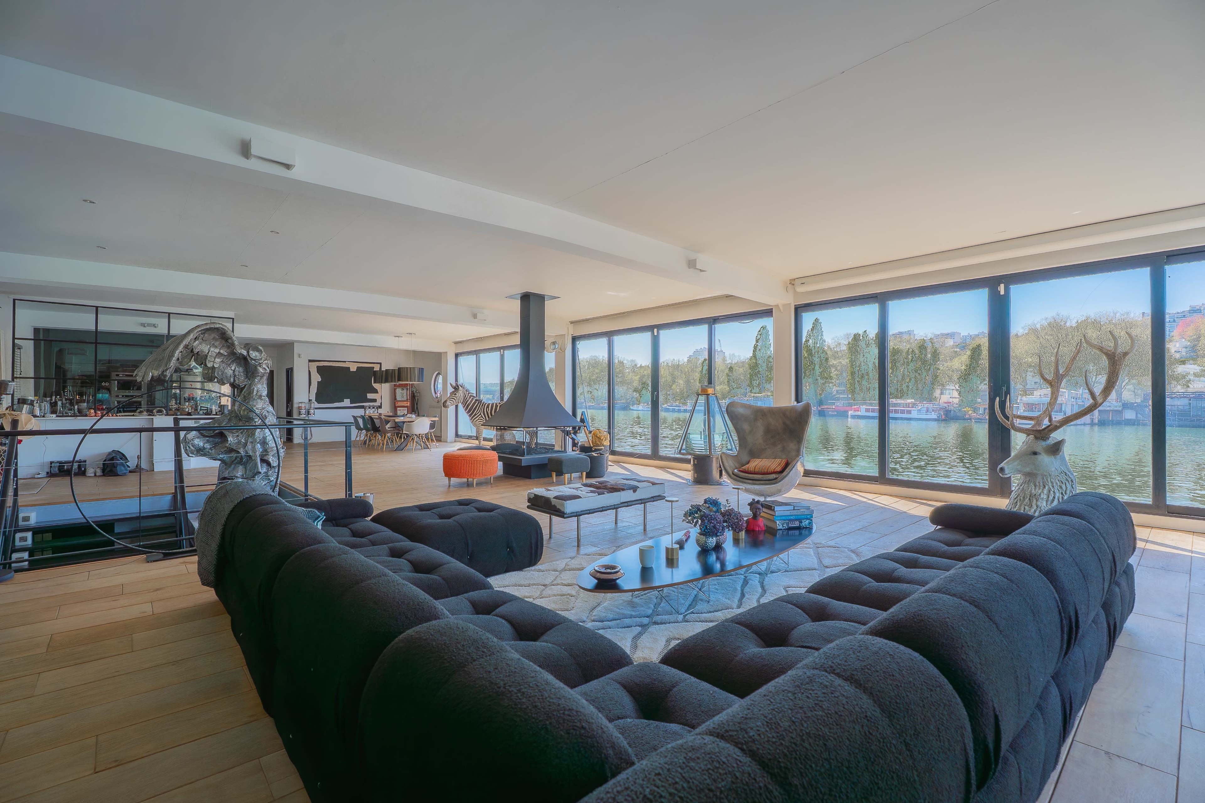 The image shows a spacious, modern living area with large windows overlooking a body of water, featuring a sectional sofa and various decorative elements.