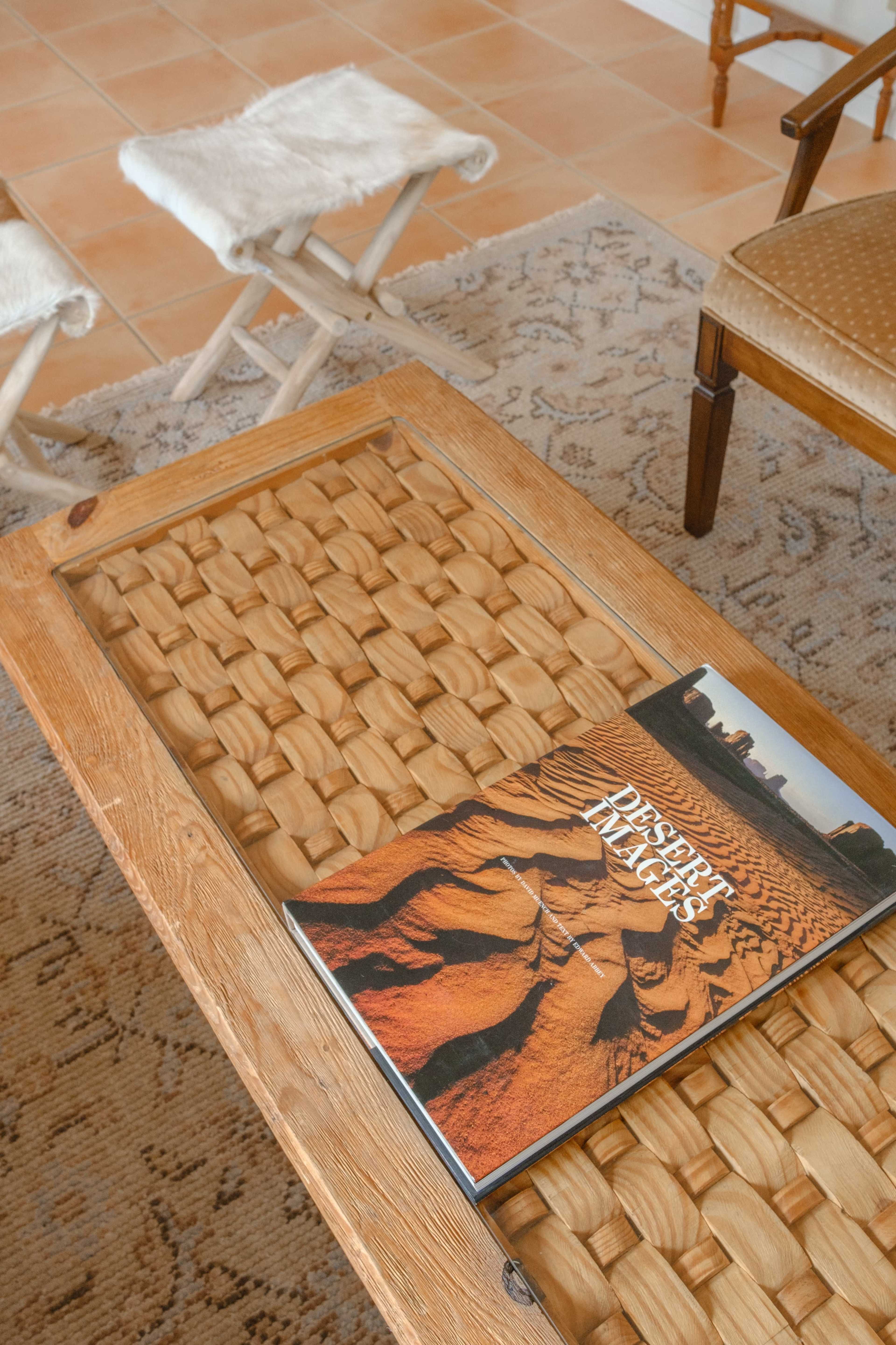 A wooden coffee table with a glass top, featuring a book titled "Desert Images" placed on a woven surface, is positioned in a room with tiled floors and wooden chairs.