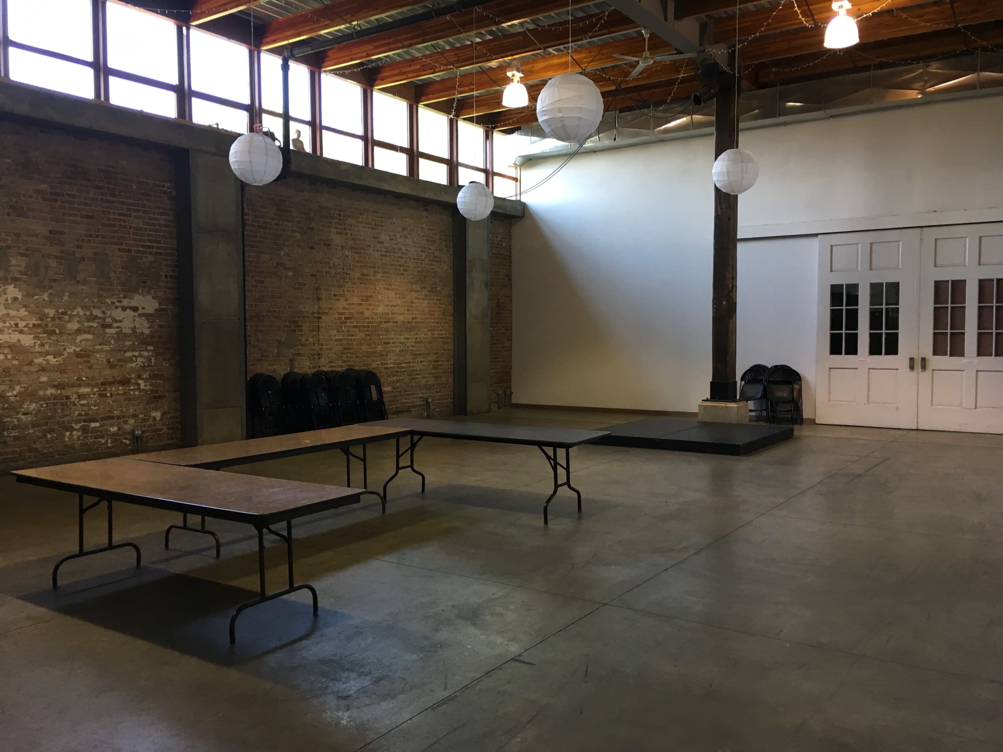 An empty room with exposed brick walls, wooden beams, and several tables arranged in the center, illuminated by hanging lanterns.