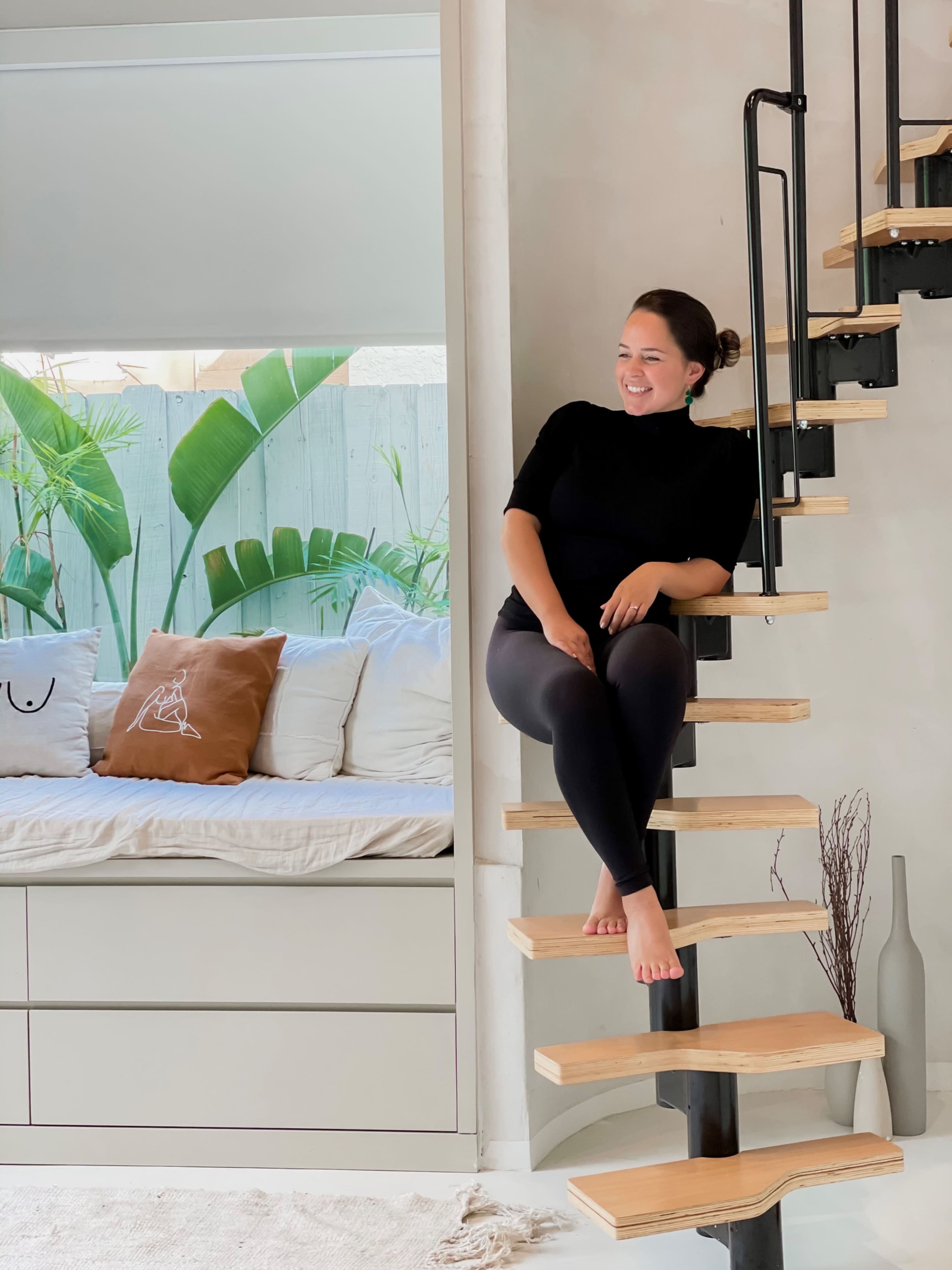 A woman sits on a wooden staircase in a modern space, beside a cozy seating area with decorative pillows and lush green plants in the background.