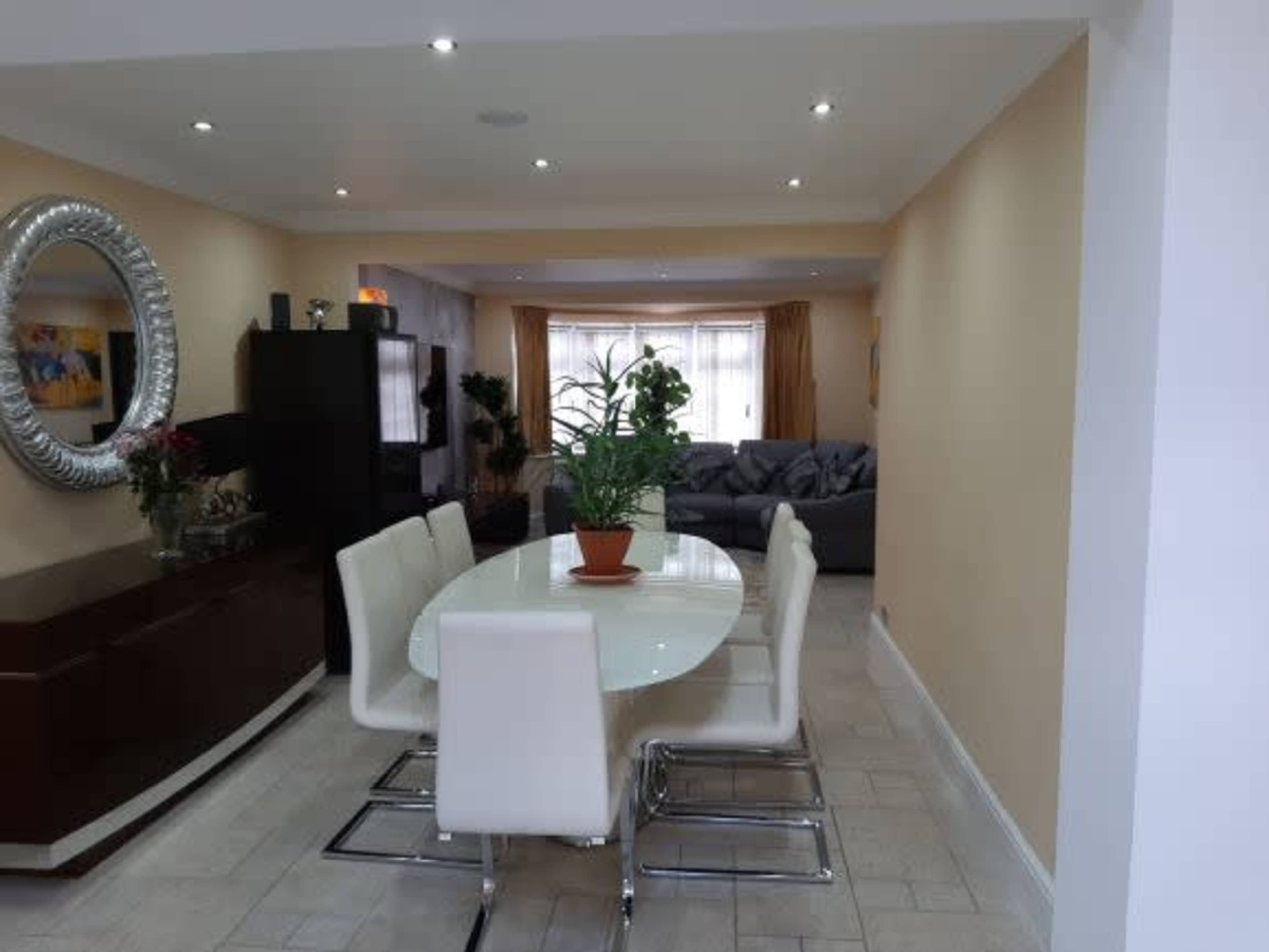 The image shows a dining area with a white oval table surrounded by six white chairs, set against a backdrop of light-colored walls and a sofa in the distance.